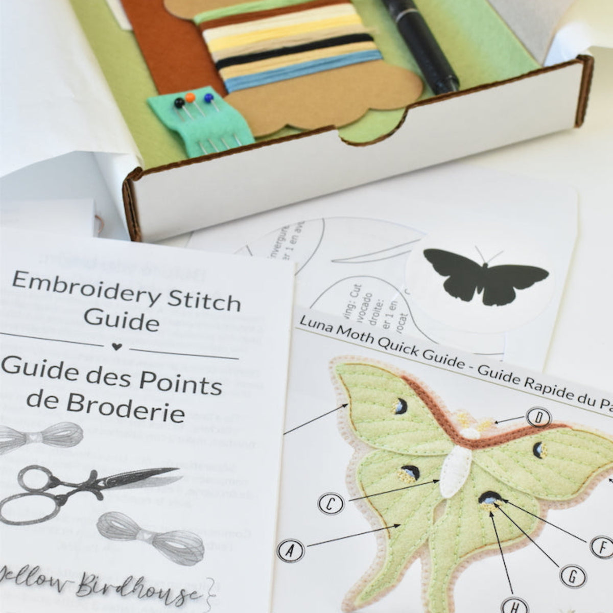 An open white and kraft cardboard box containing embroidery starter supplies, including a black pen, ribbon, thread, and a small turquoise item, alongside printed guides titled 'Embroidery Stitch Guide' and 'Luna Moth Quick Guide' on a white surface.