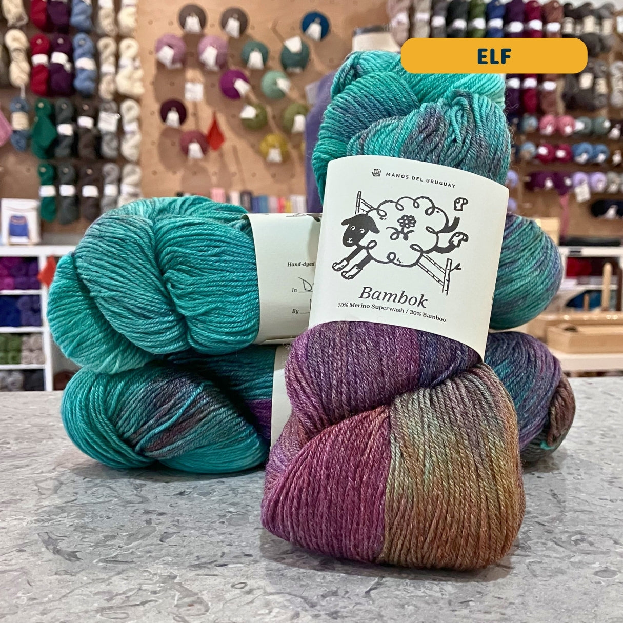 100g multi-coloured teal, purple, orange variegated (elf) merino bamboo sock yarn, 'Manos del Uruguay Bambok 70% Merino Superwash / 30% Bamboo' label visible, craft shop background (blurred)

