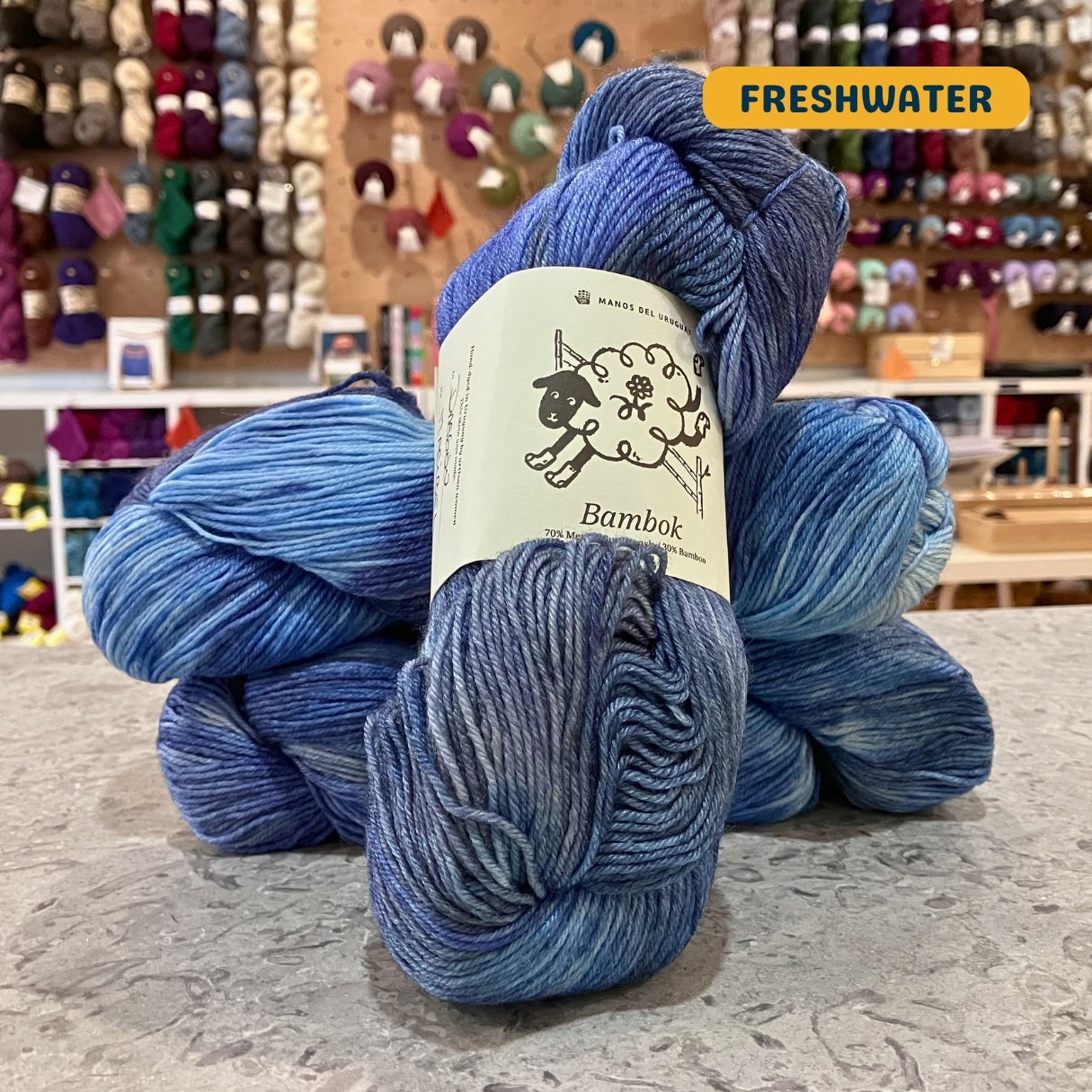 100g blue variegated (freshwater) merino bamboo sock yarn, 'Manos del Uruguay Bambok 70% Merino Superwash / 30% Bamboo' label visible, craft shop background (blurred)
