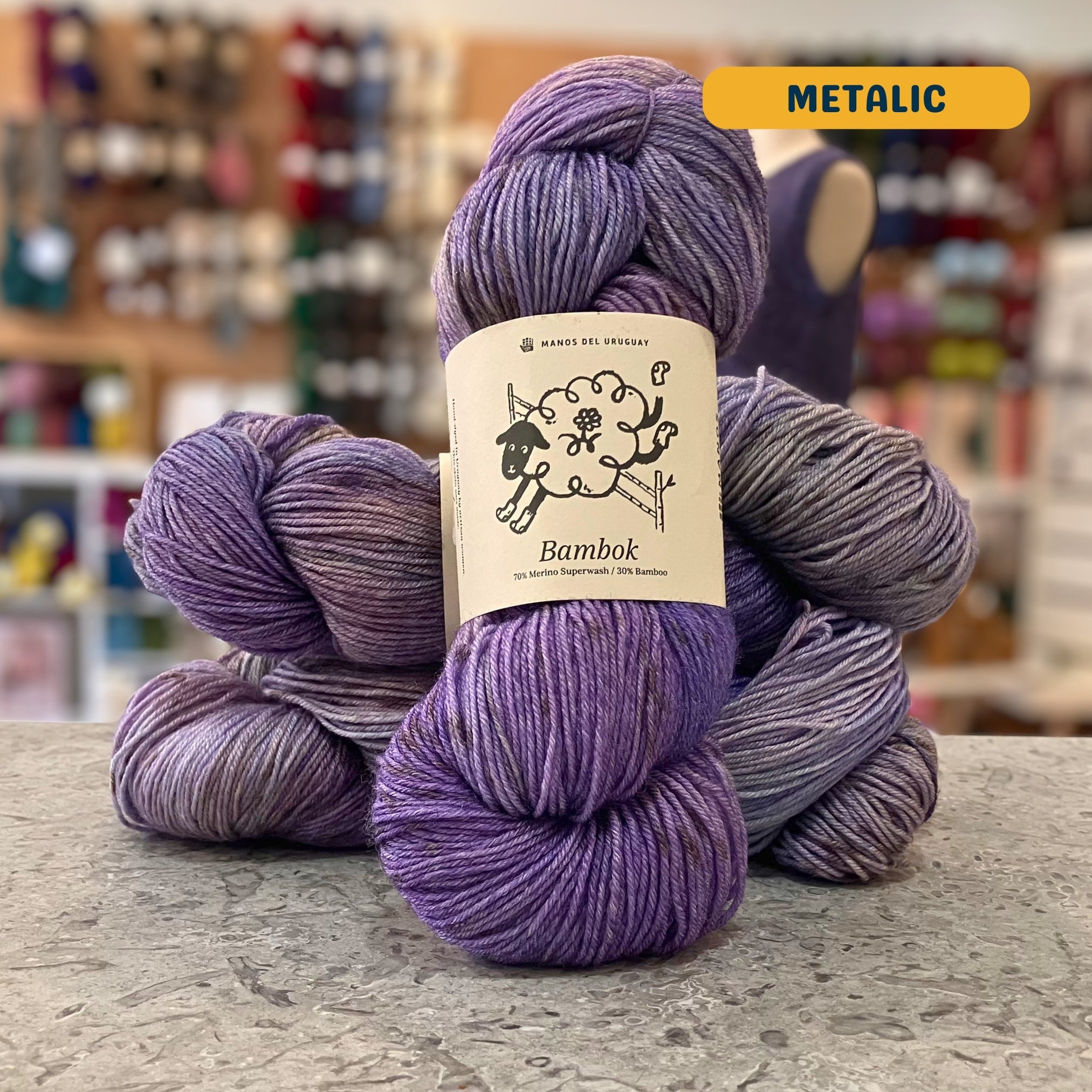 100g purple variegated (metalic) merino bamboo sock yarn, 'Manos del Uruguay Bambok 70% Merino Superwash / 30% Bamboo' label visible, craft shop background (blurred)
