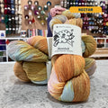 100g multi-coloured orange, pink, blue variegated (nectar) merino bamboo sock yarn, 'Manos del Uruguay Bambok 70% Merino Superwash / 30% Bamboo' label visible, craft shop background (blurred)

