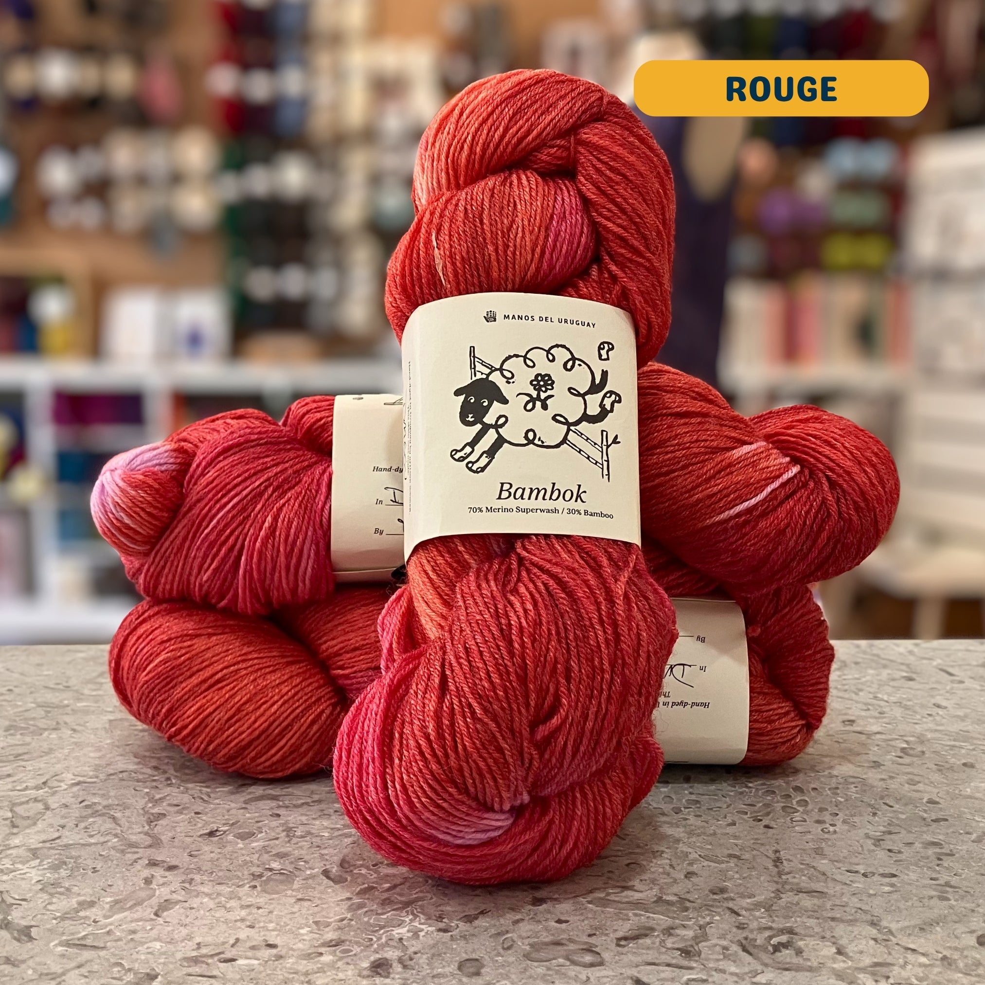100g red & pink variegated (rouge) merino bamboo sock yarn, 'Manos del Uruguay Bambok 70% Merino Superwash / 30% Bamboo' label visible, craft shop background (blurred)
