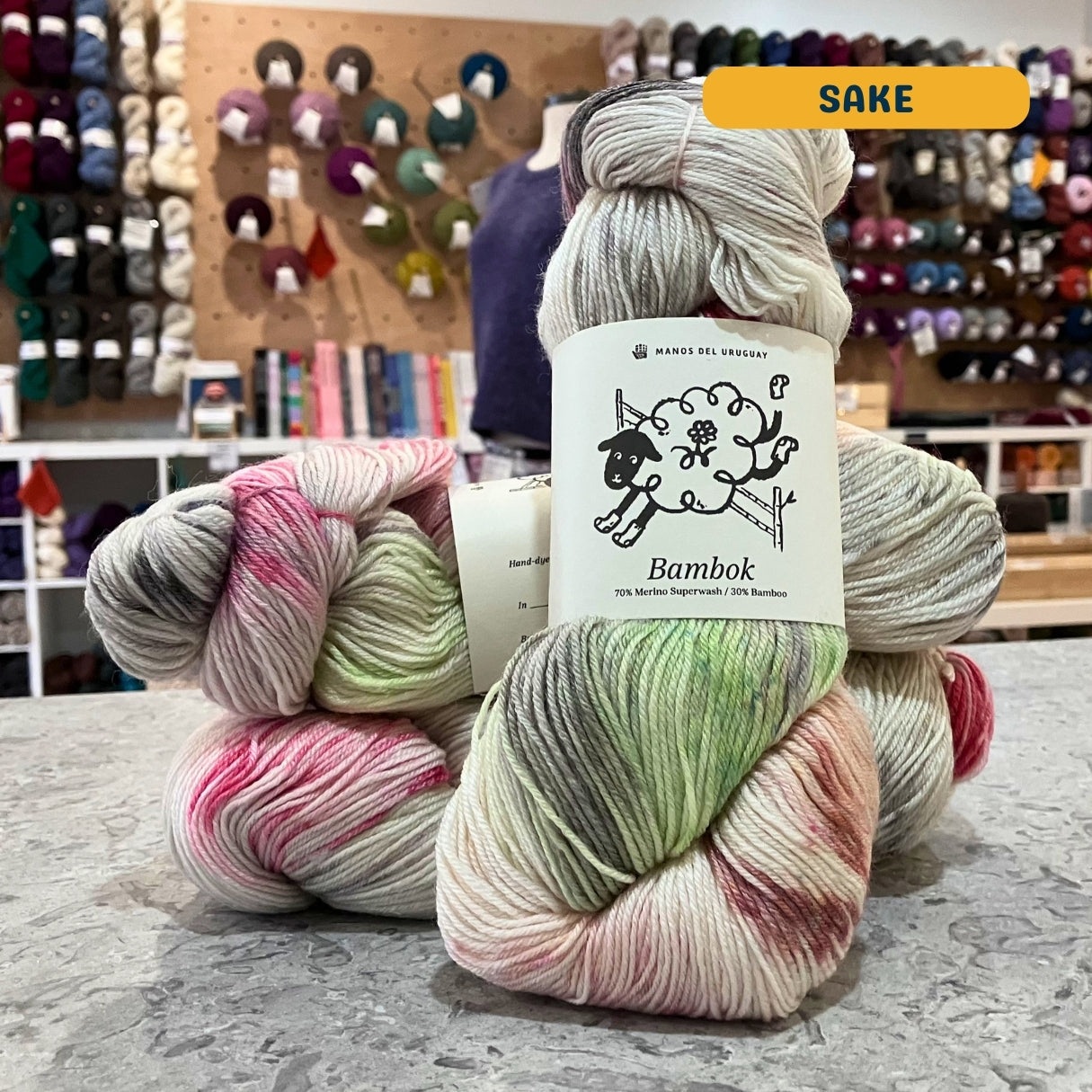 100g multi-coloured light grey, green, pink variegated (sake) merino bamboo sock yarn, 'Manos del Uruguay Bambok 70% Merino Superwash / 30% Bamboo' label visible, craft shop background (blurred)