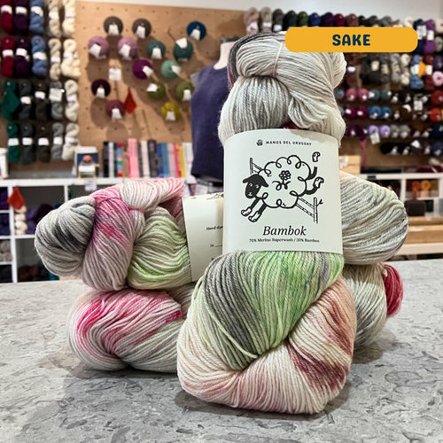 100g multi-coloured light grey, green, pink variegated (sake) merino bamboo sock yarn, 'Manos del Uruguay Bambok 70% Merino Superwash / 30% Bamboo' label visible, craft shop background (blurred)