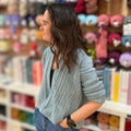 A woman with long dark hair is shown in profile wearing a light blue-gray textured knit cardigan, standing in a yarn or fabric store with shelves of colorful yarn balls blurred in the background.