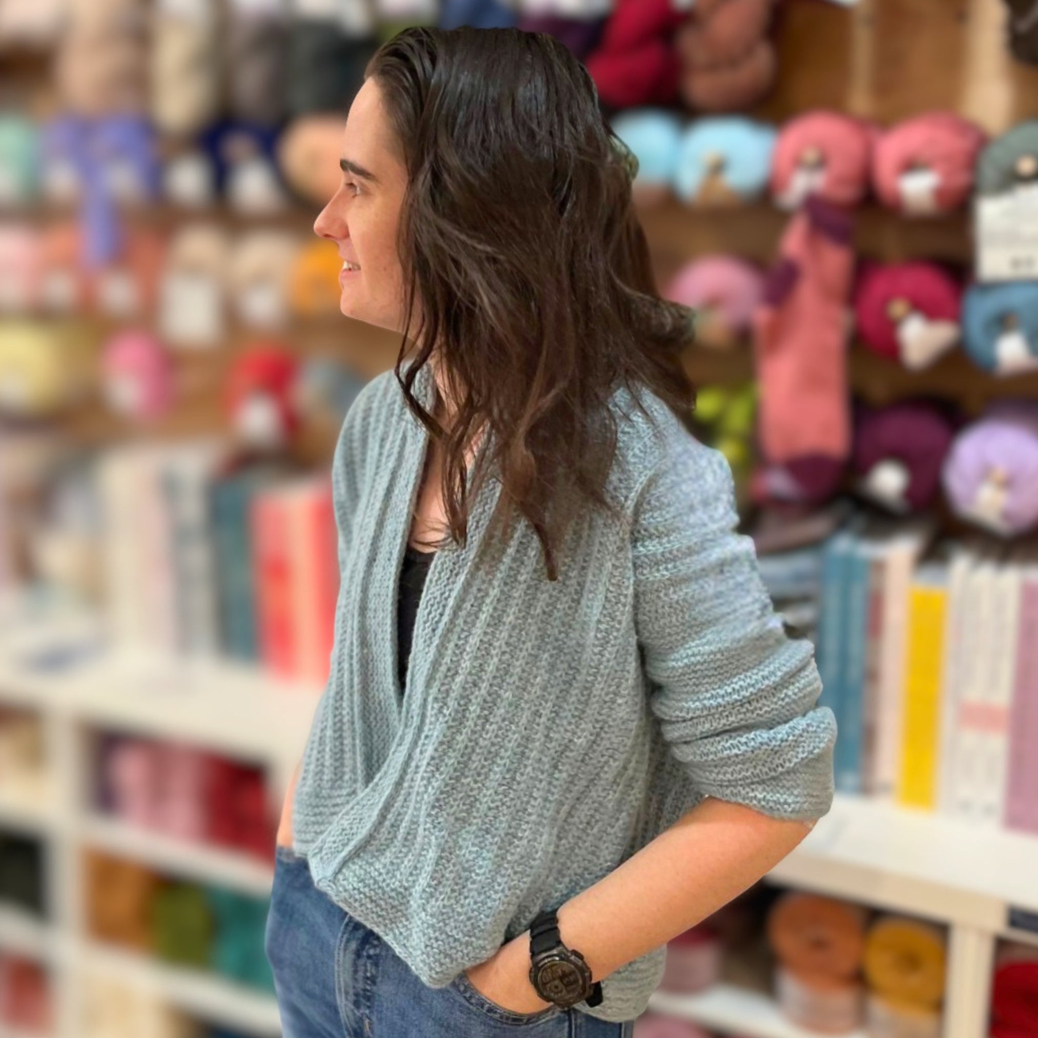 A woman with long dark hair is shown in profile wearing a light blue-gray textured knit cardigan, standing in a yarn or fabric store with shelves of colorful yarn balls blurred in the background.