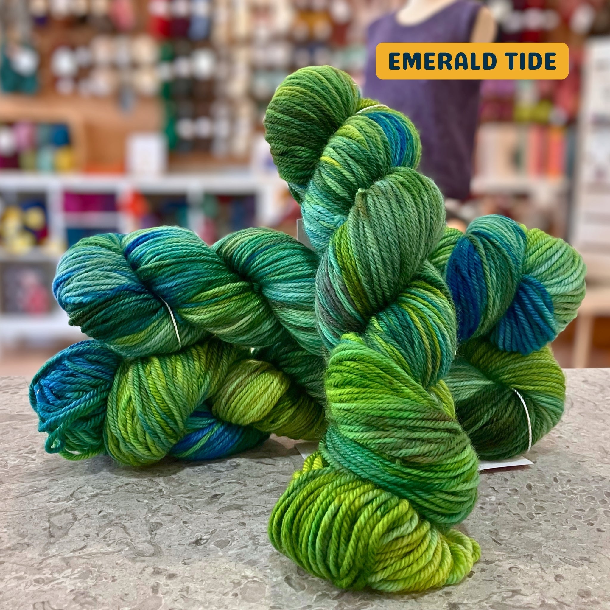 100g variegated blue, teal, green (emerald tide) hand-dyed SW merino worsted yarn, 'Nova Knits Worsted', craft shop background (blurred)