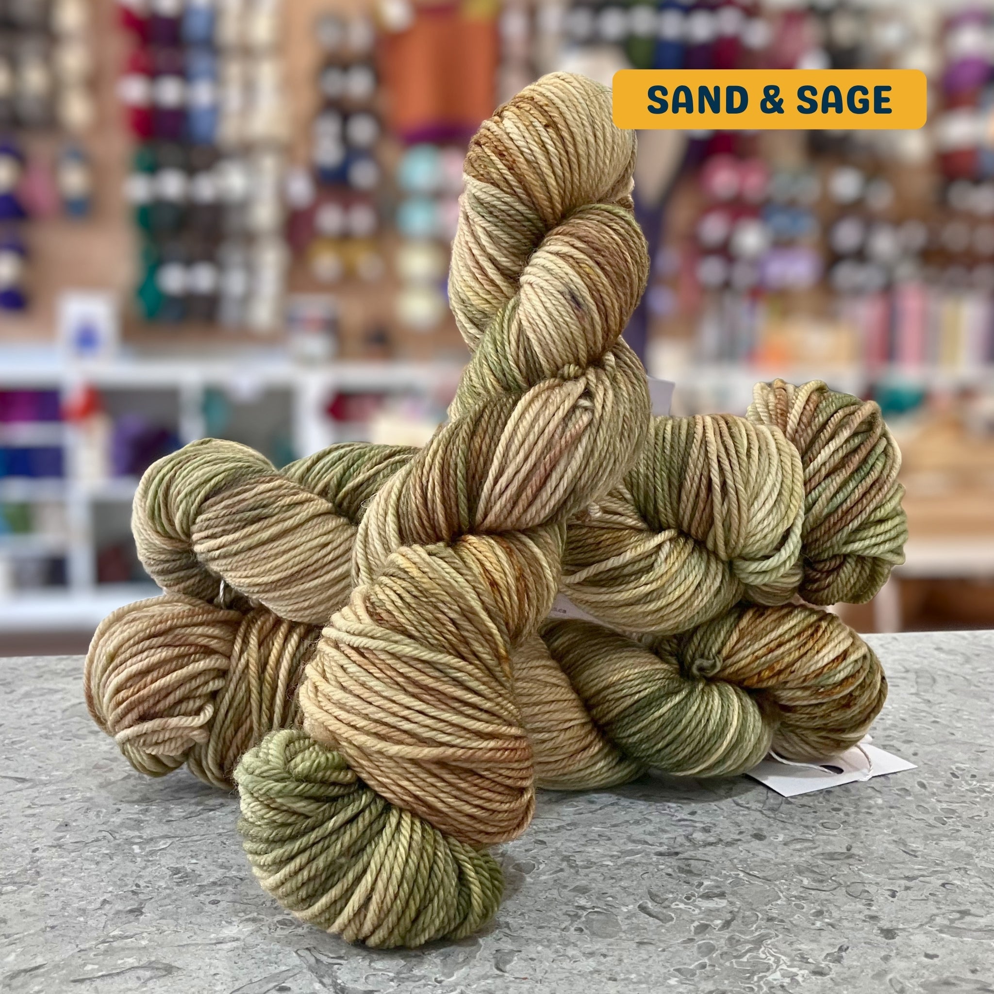 100g variegated brown, green (sand & sage) hand-dyed SW merino worsted yarn, 'Nova Knits Worsted', craft shop background (blurred)