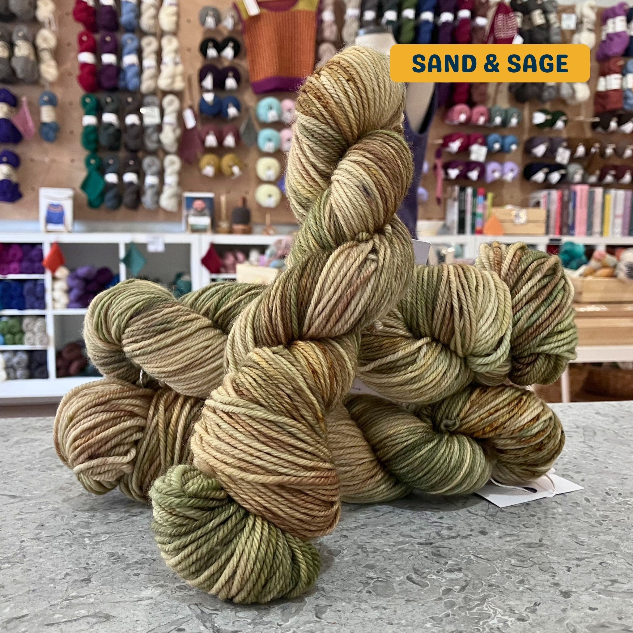 100g variegated brown, green (sand & sage) hand-dyed SW merino worsted yarn, 'Nova Knits Worsted', craft shop background (blurred)