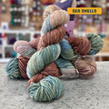 100g variegated pink, blue, green (sea shells) hand-dyed SW merino worsted yarn, 'Nova Knits Worsted', craft shop background (blurred)
