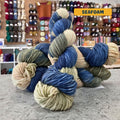 100g variegated blue, pink-beige, green-grey (seafoam) hand-dyed SW merino worsted yarn, 'Nova Knits Worsted', craft shop background (blurred)