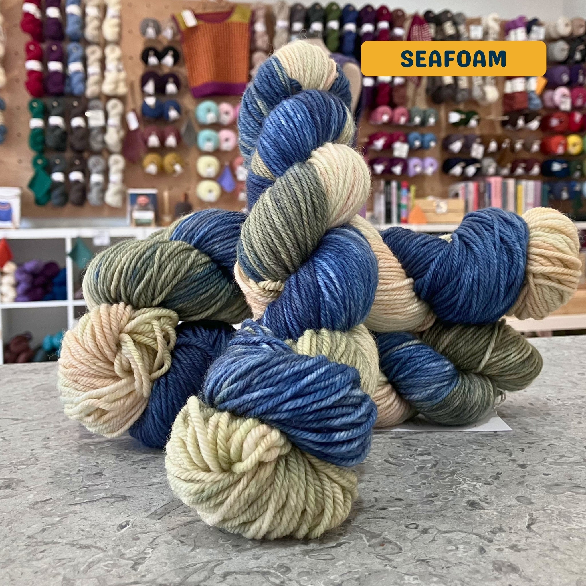 100g variegated blue, pink-beige, green-grey (seafoam) hand-dyed SW merino worsted yarn, 'Nova Knits Worsted', craft shop background (blurred)