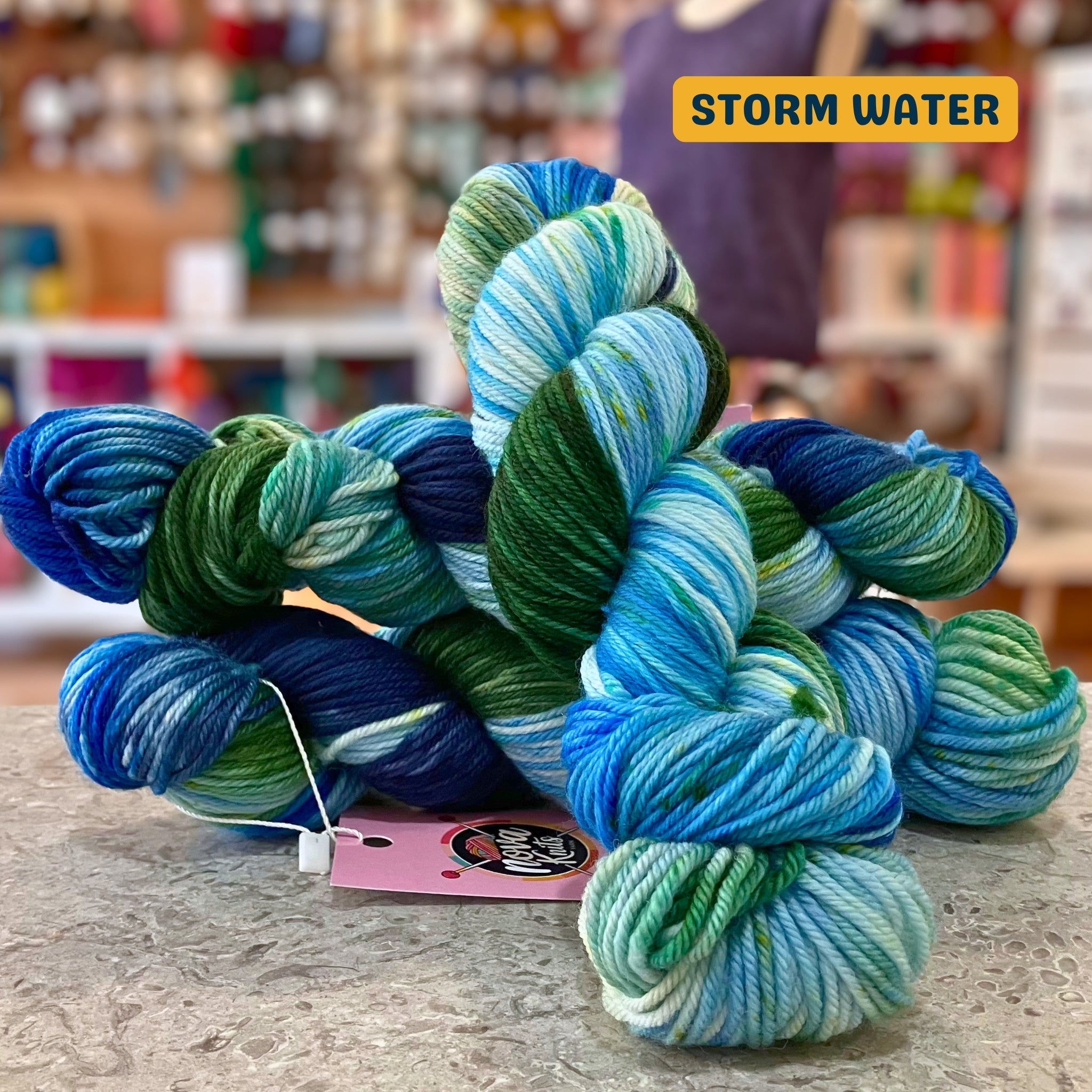 100g variegated blue, green (storm water) hand-dyed SW merino worsted yarn, 'Nova Knits Worsted', craft shop background (blurred)