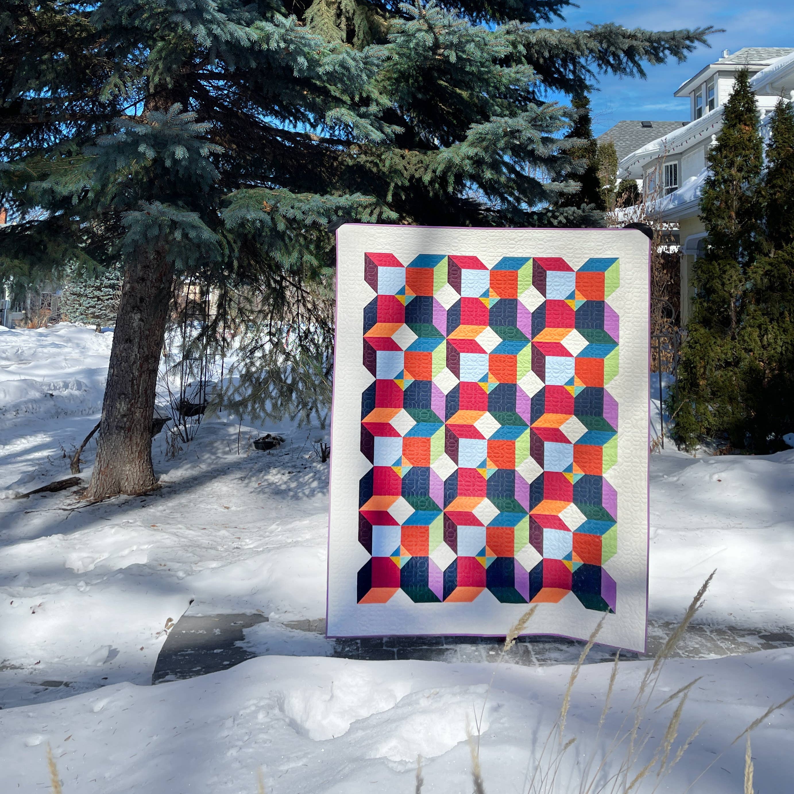 Colourful geometric quilt displayed outdoors in a snowy landscape with trees.