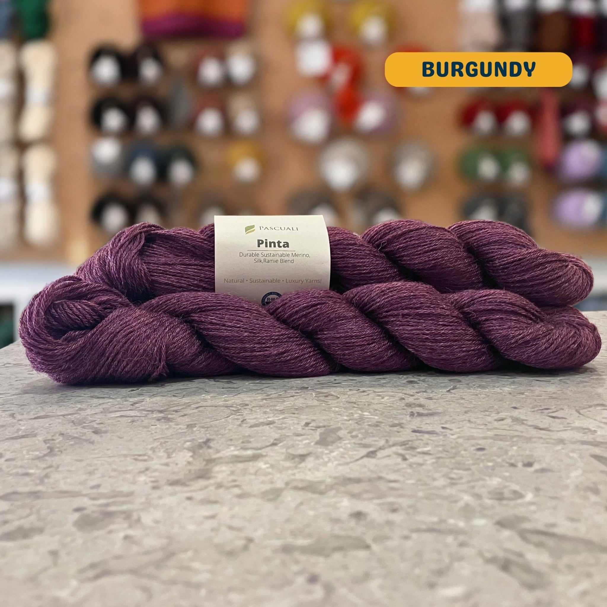 50g dark purple (burgundy) merino, silk, & ramie sock yarn, 'Pascuali Pinta Durable Organic Merino Silk Ramie Blend' label visible, craft shop background (blurred)