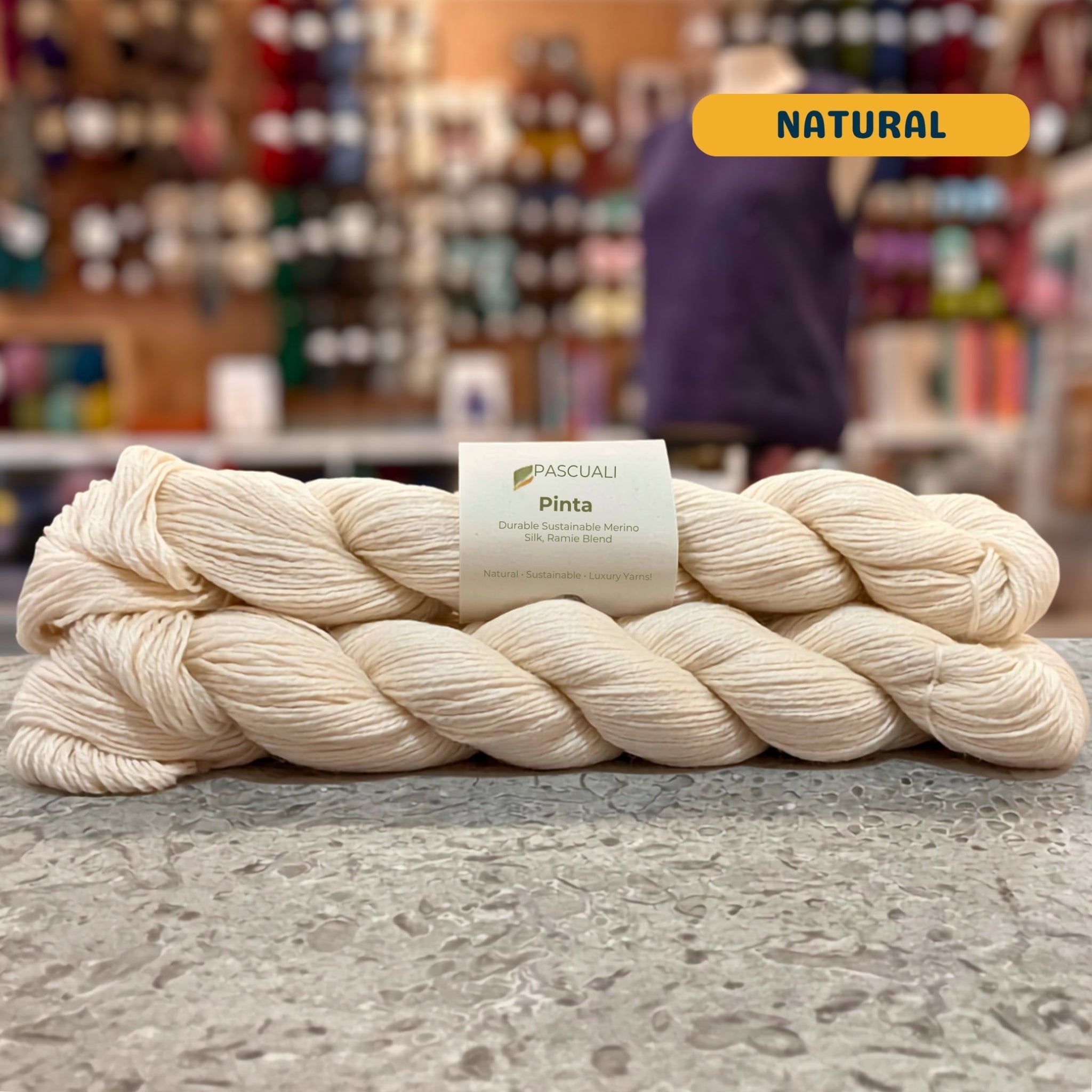 50g ecru (natural) merino, silk, & ramie sock yarn, 'Pascuali Pinta Durable Organic Merino Silk Ramie Blend' label visible, craft shop background (blurred)
