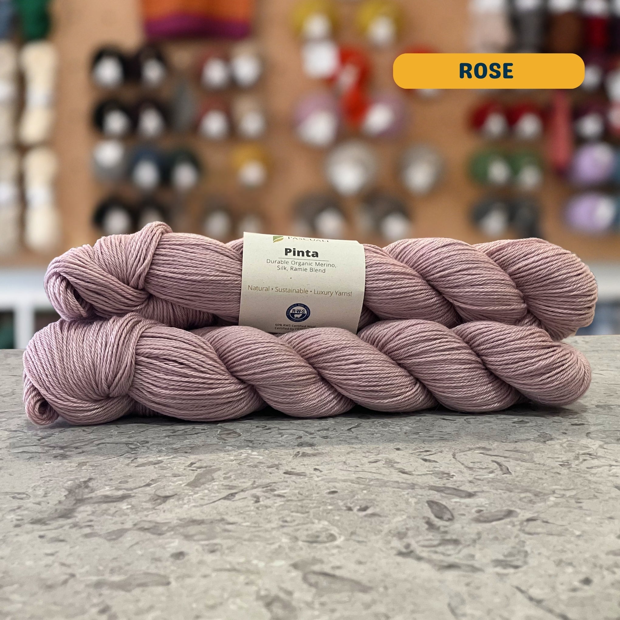 50g light grey-pink (rose) merino, silk, & ramie sock yarn, 'Pascuali Pinta Durable Organic Merino Silk Ramie Blend' label visible, craft shop background (blurred)