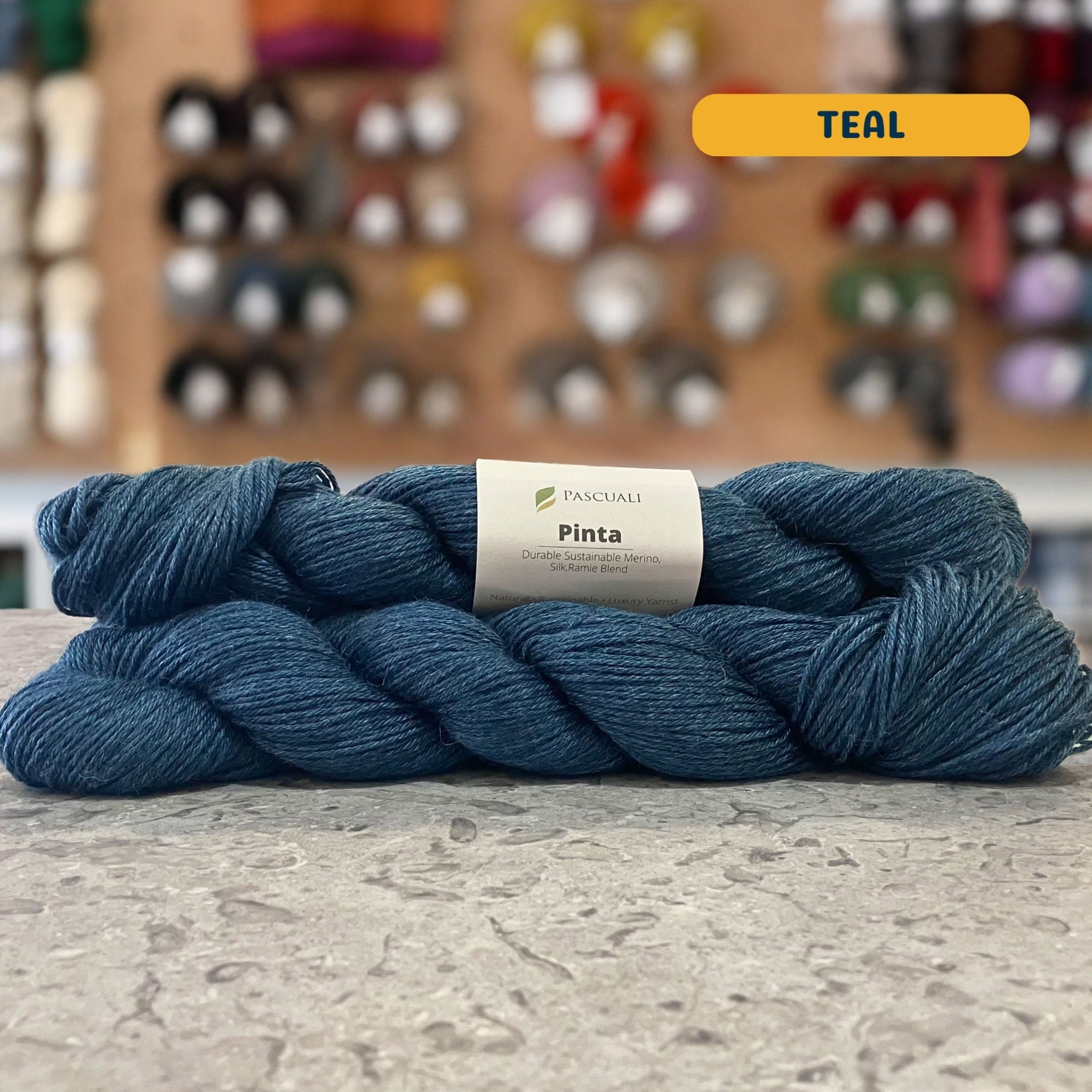 50g dark blue (teal) merino, silk, & ramie sock yarn, 'Pascuali Pinta Durable Organic Merino Silk Ramie Blend' label visible, craft shop background (blurred)