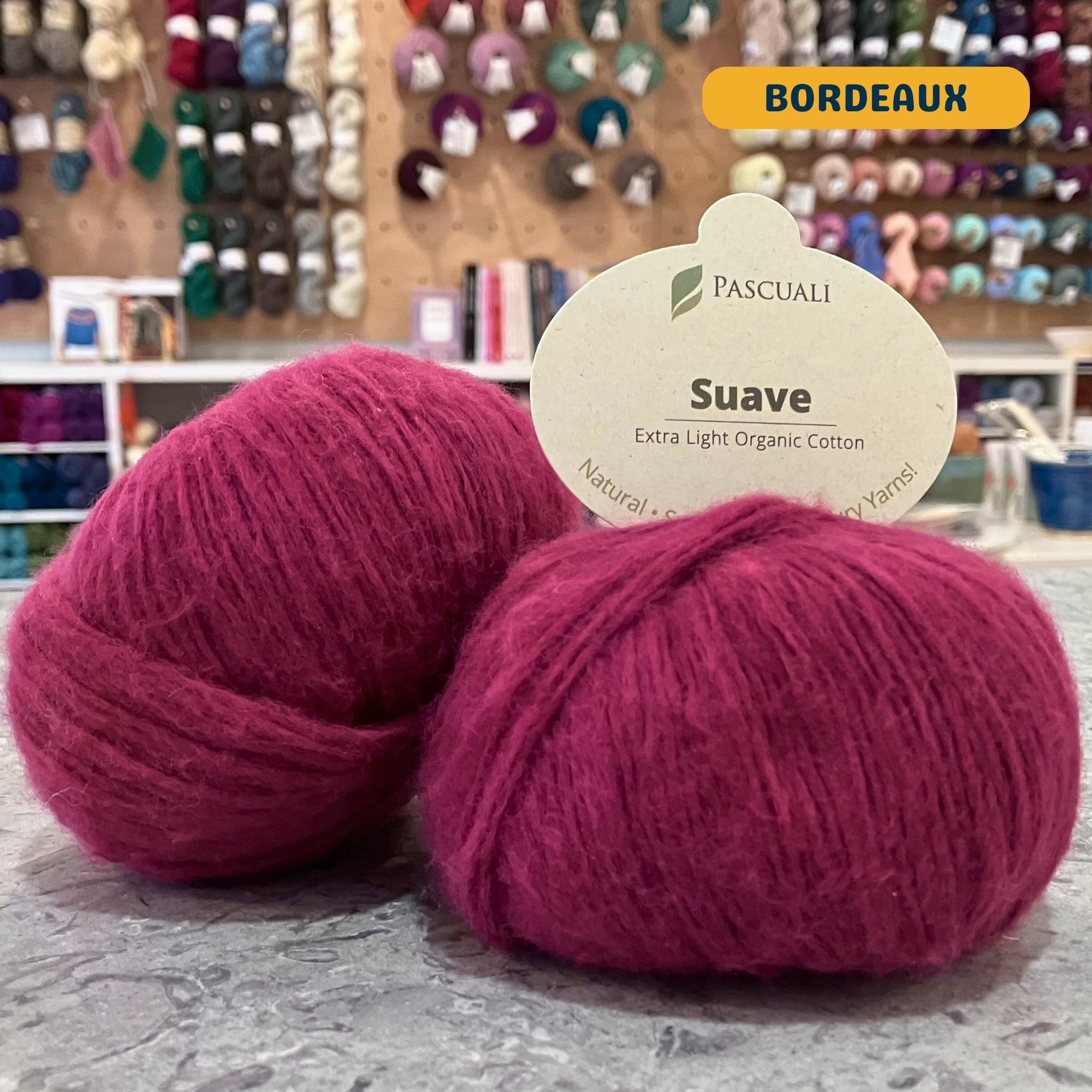 25g red (bordeaux) organic cotton sport yarn, 'Pascuali Suave Extra Light Organic Cotton' label visible, craft shop background (blurred)