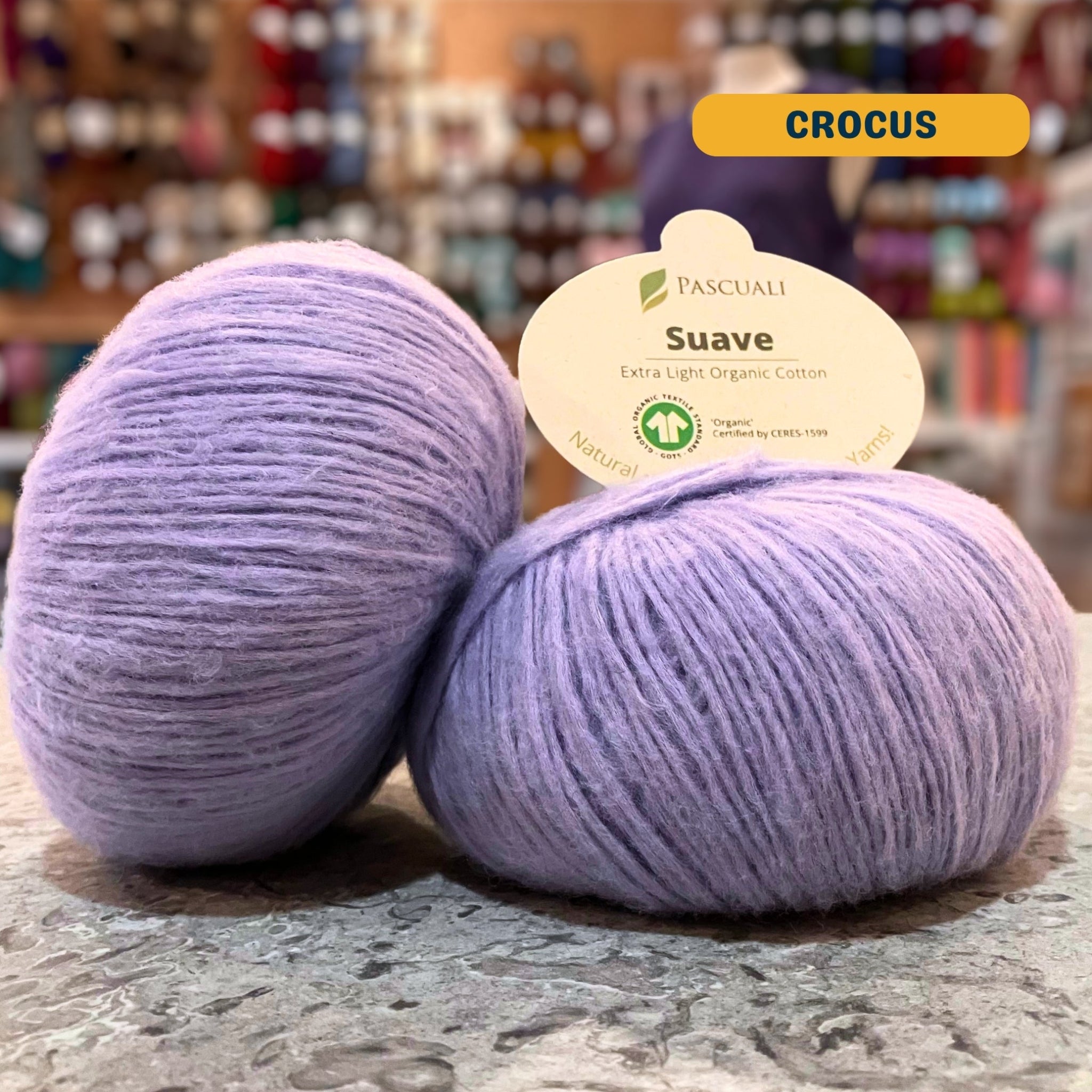 25g light purple (crocus) organic cotton sport yarn, 'Pascuali Suave Extra Light Organic Cotton' label visible, craft shop background (blurred)