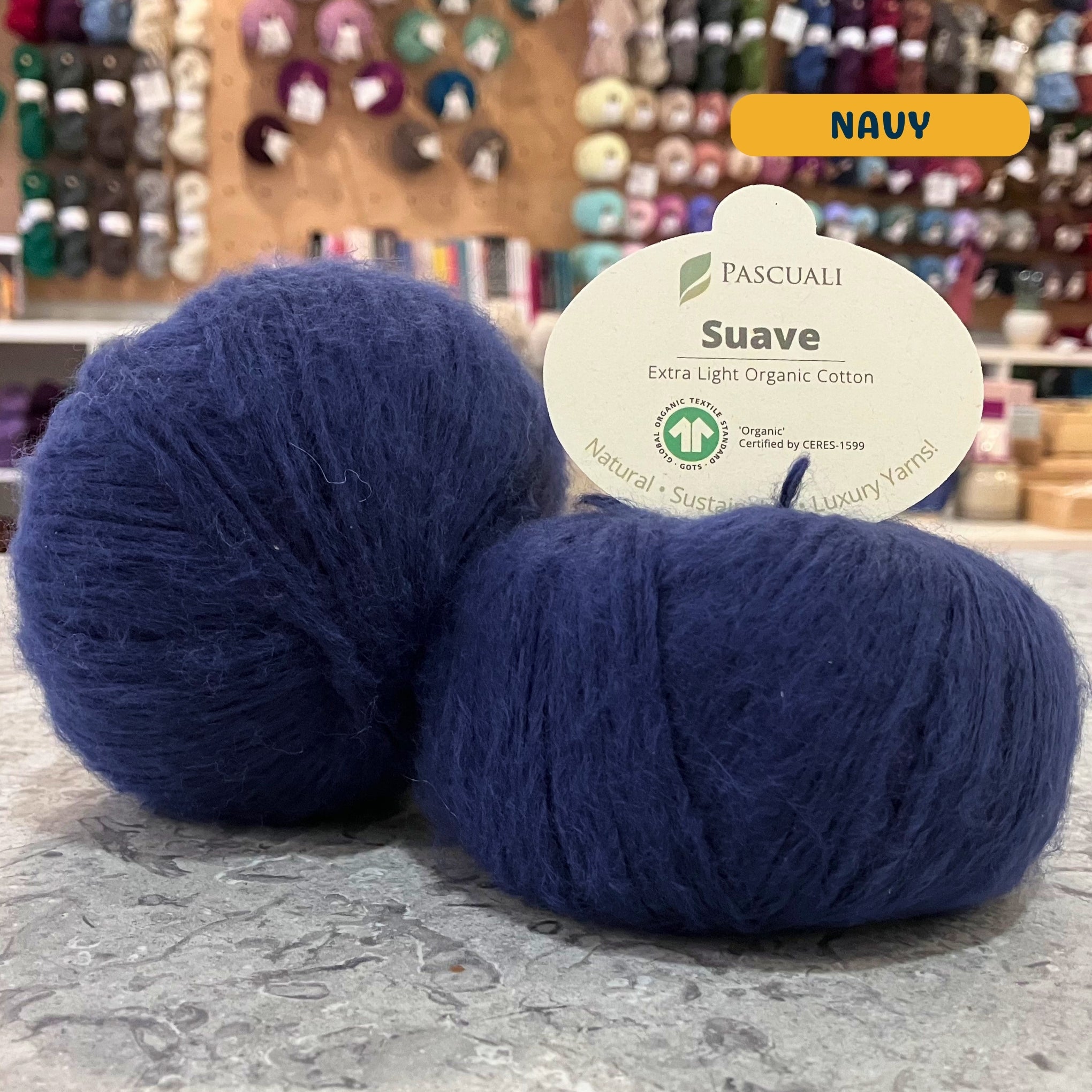 25g navy blue organic cotton sport yarn, 'Pascuali Suave Extra Light Organic Cotton' label visible, craft shop background (blurred)