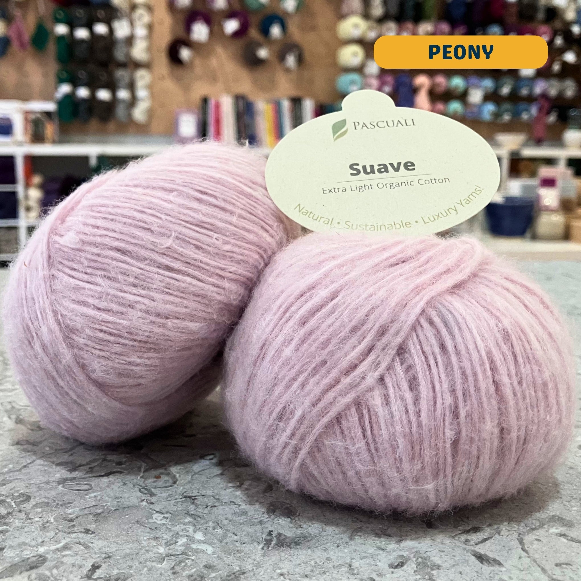 25g pale pink (peony) organic cotton sport yarn, 'Pascuali Suave Extra Light Organic Cotton' label visible, craft shop background (blurred)
