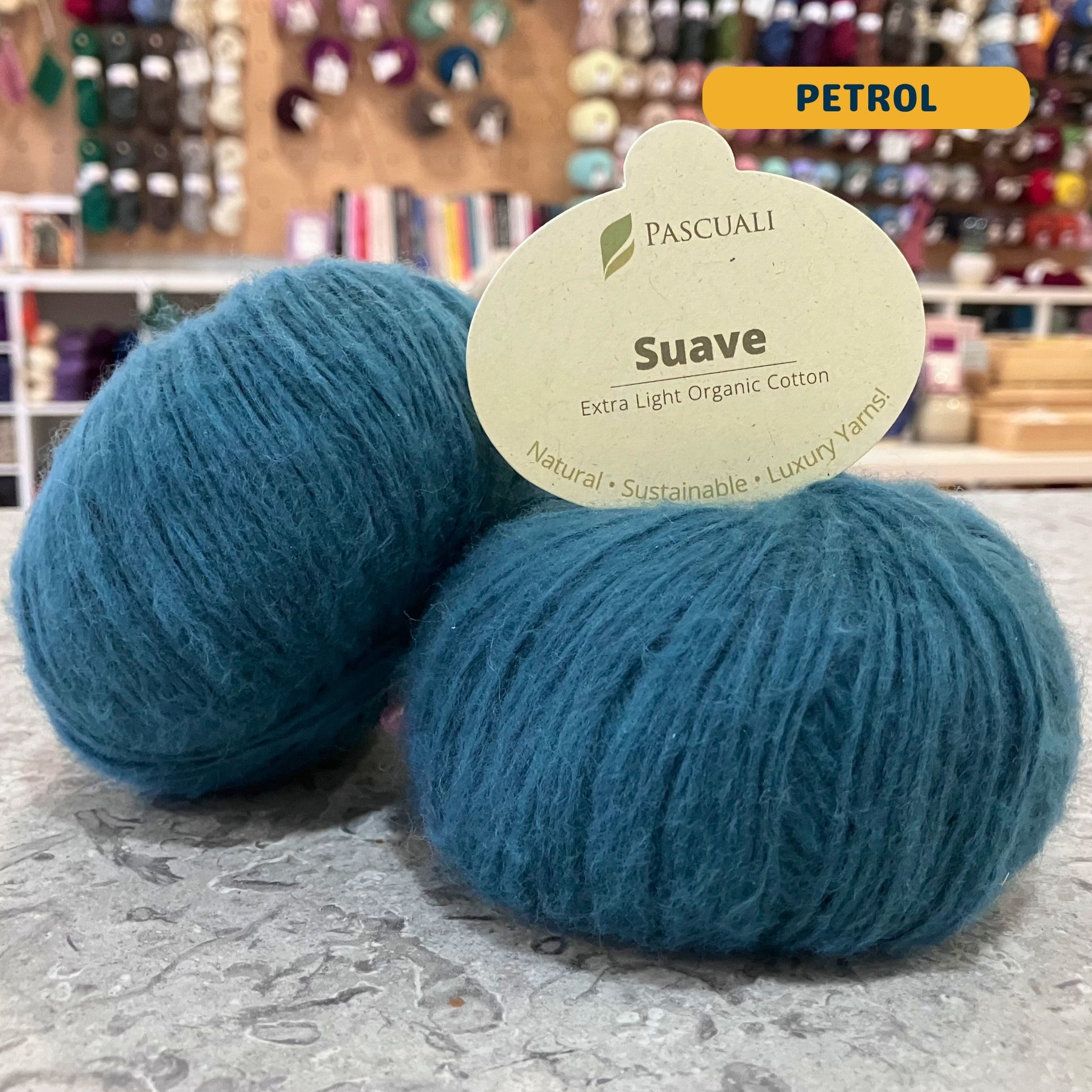 25g teal (petrol) organic cotton sport yarn, 'Pascuali Suave Extra Light Organic Cotton' label visible, craft shop background (blurred)