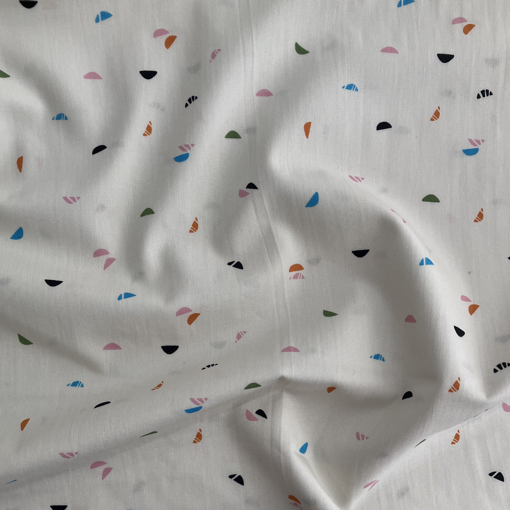 100% cotton quilting fabric — light cream fabric with green, blue, pink, orange, and black crescent shapes. 44/45in width.