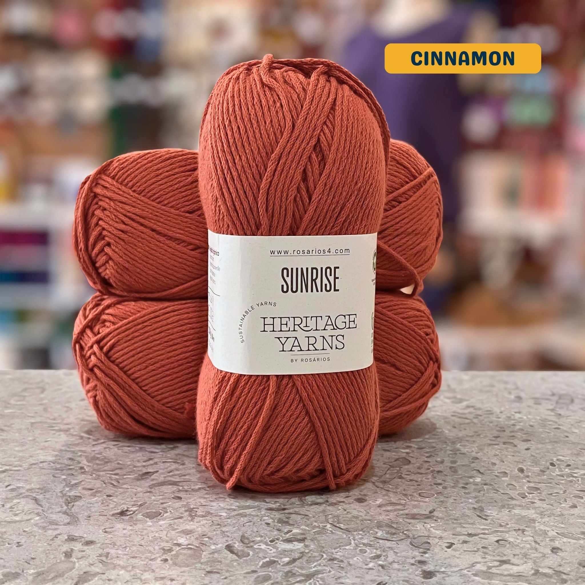 100g orange-brown (cinnamon) organic cotton worsted yarn, 'Sunrise Sustainable Yarns Heritage Yarns by Rosarios' label visible, craft shop background (blurred)