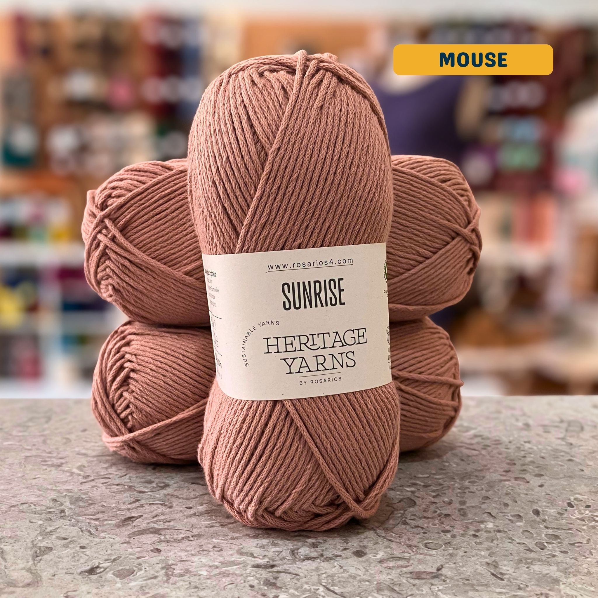 100g light pink-brown (mouse) organic cotton worsted yarn, 'Sunrise Sustainable Yarns Heritage Yarns by Rosarios' label visible, craft shop background (blurred)