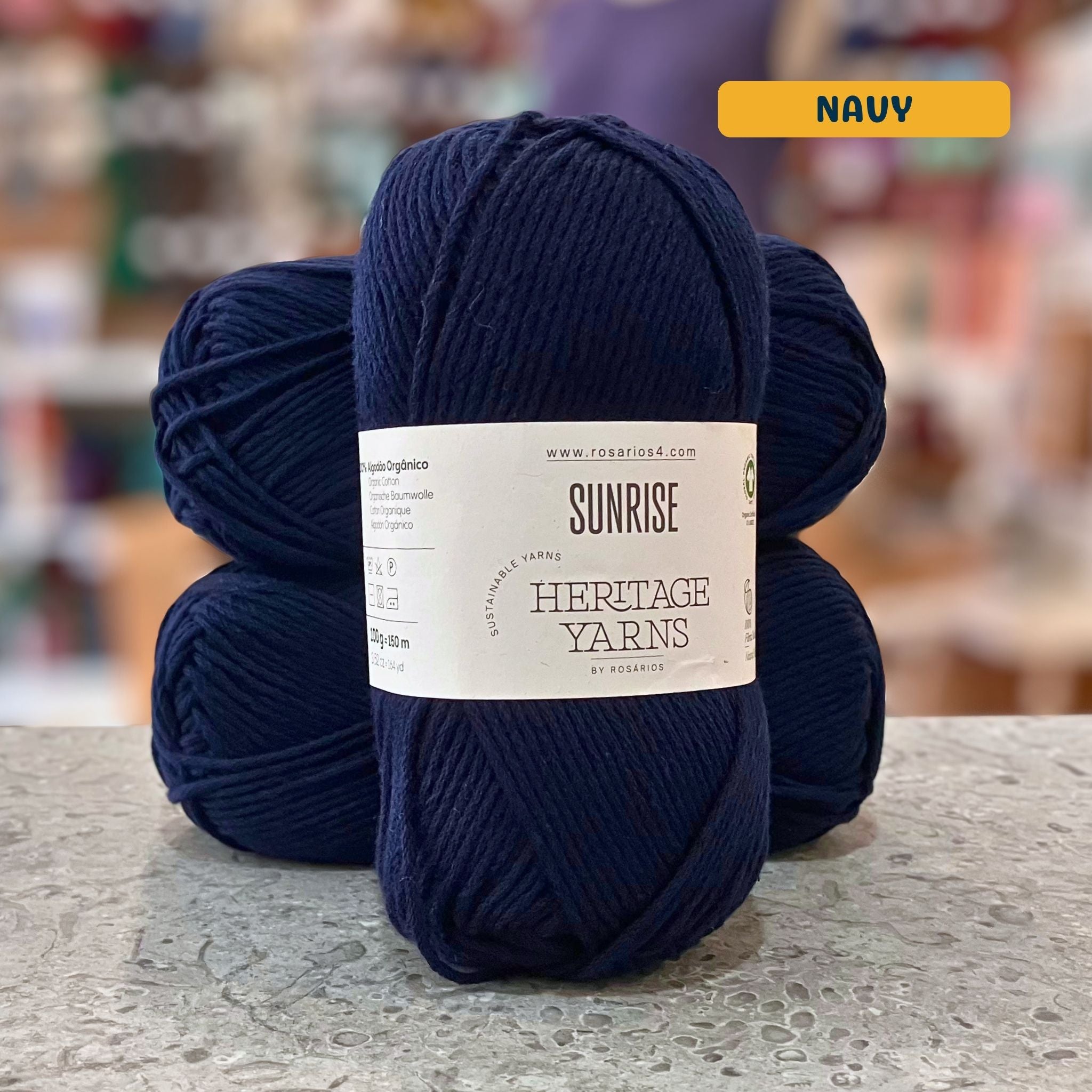 100g dark blue (navy) organic cotton worsted yarn, 'Sunrise Sustainable Yarns Heritage Yarns by Rosarios' label visible, craft shop background (blurred)
