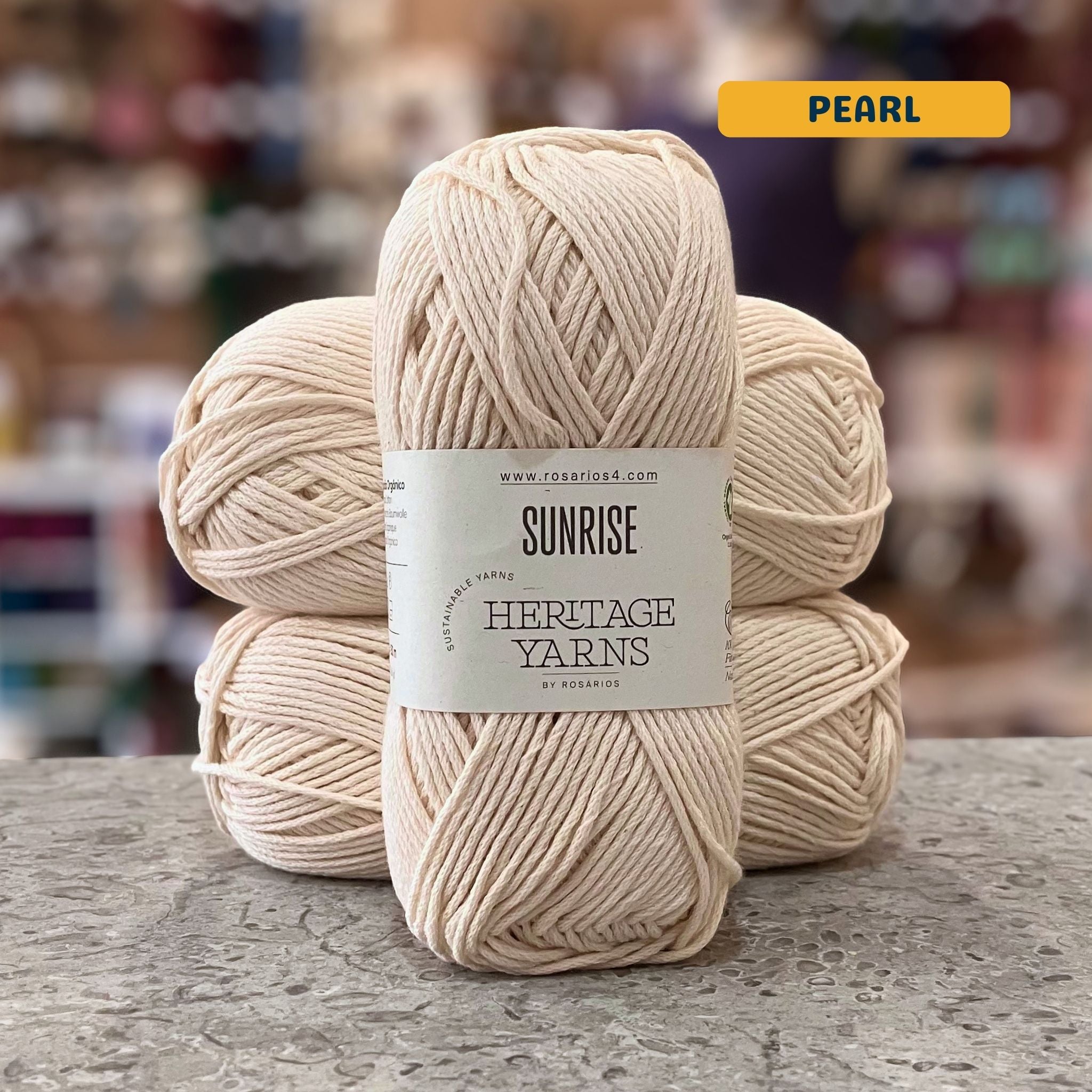 100g natural white (pearl) organic cotton worsted yarn, 'Sunrise Sustainable Yarns Heritage Yarns by Rosarios' label visible, craft shop background (blurred)