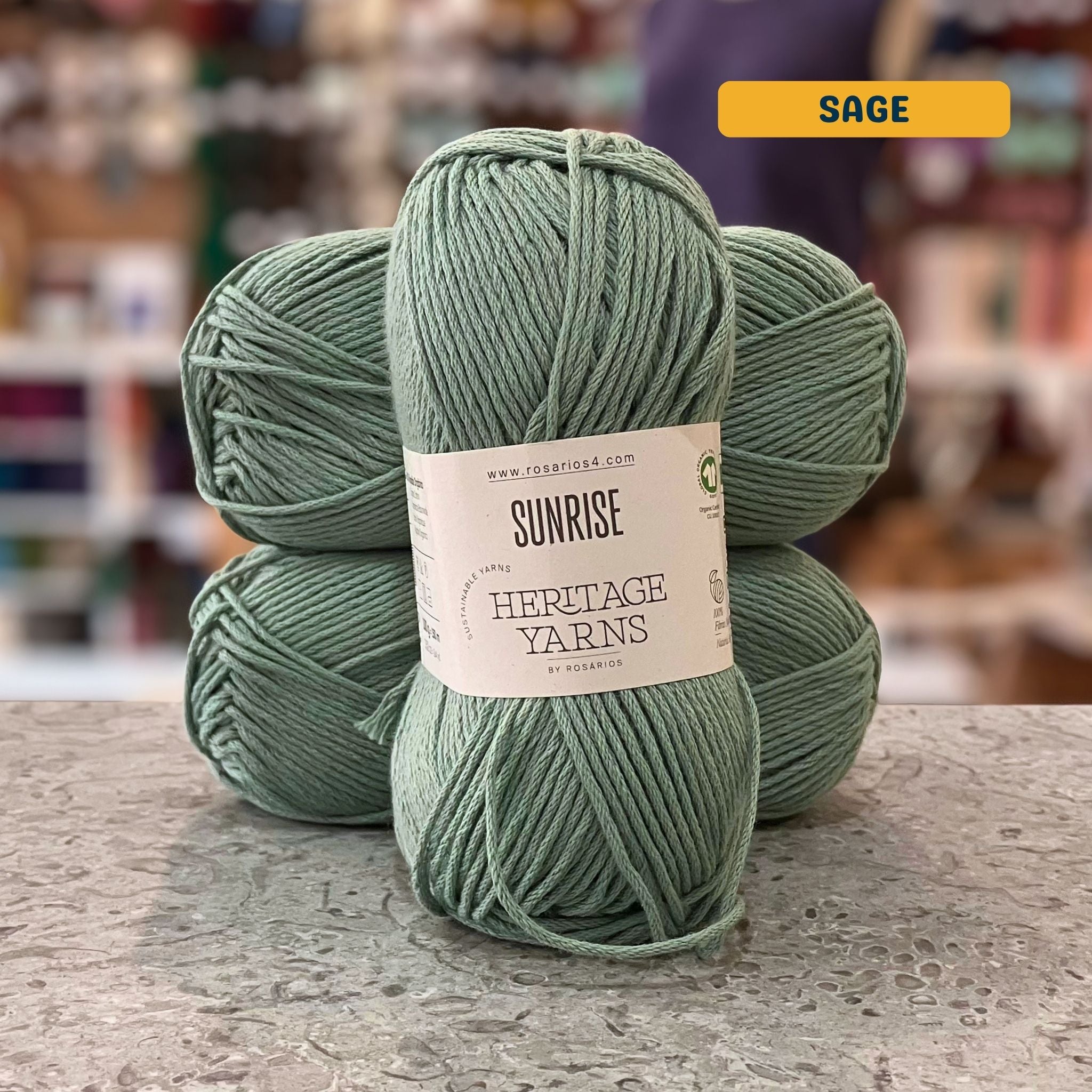 100g muted blue-green (sage) organic cotton worsted yarn, 'Sunrise Sustainable Yarns Heritage Yarns by Rosarios' label visible, craft shop background (blurred)