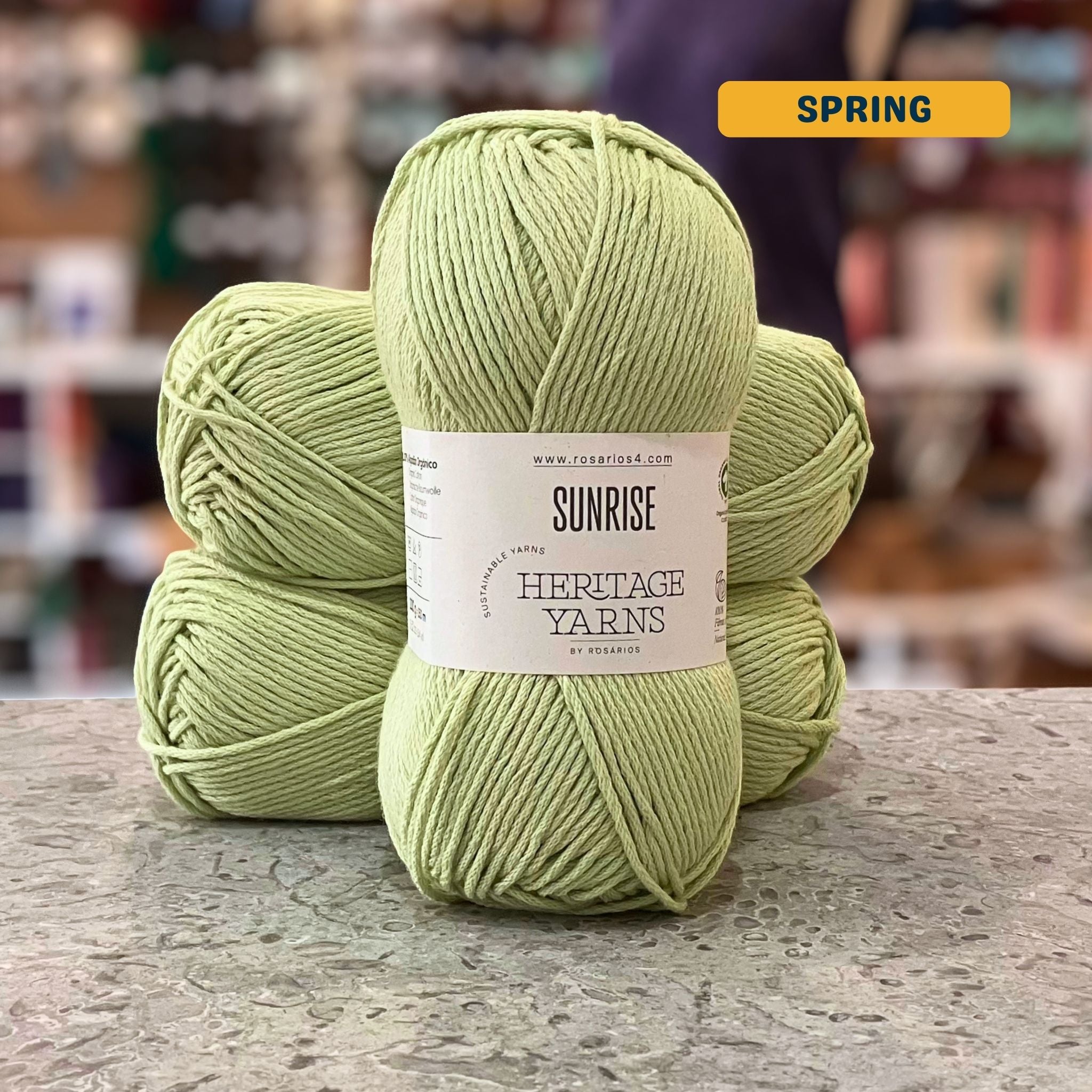 100g light green (spring) organic cotton worsted yarn, 'Sunrise Sustainable Yarns Heritage Yarns by Rosarios' label visible, craft shop background (blurred)