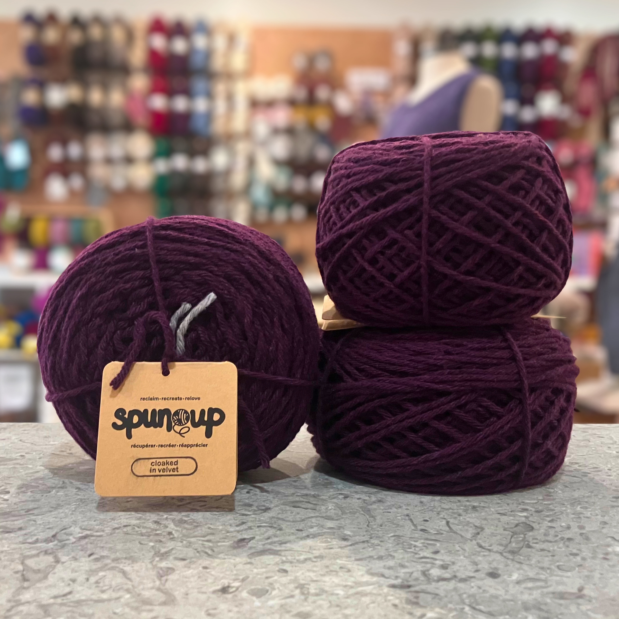 50g deep purple (cloaked in velvet) GRS-certified recycled wool worsted yarn, 'Spun Up reclaim recreate relove' label visible, craft shop background (blurred)
