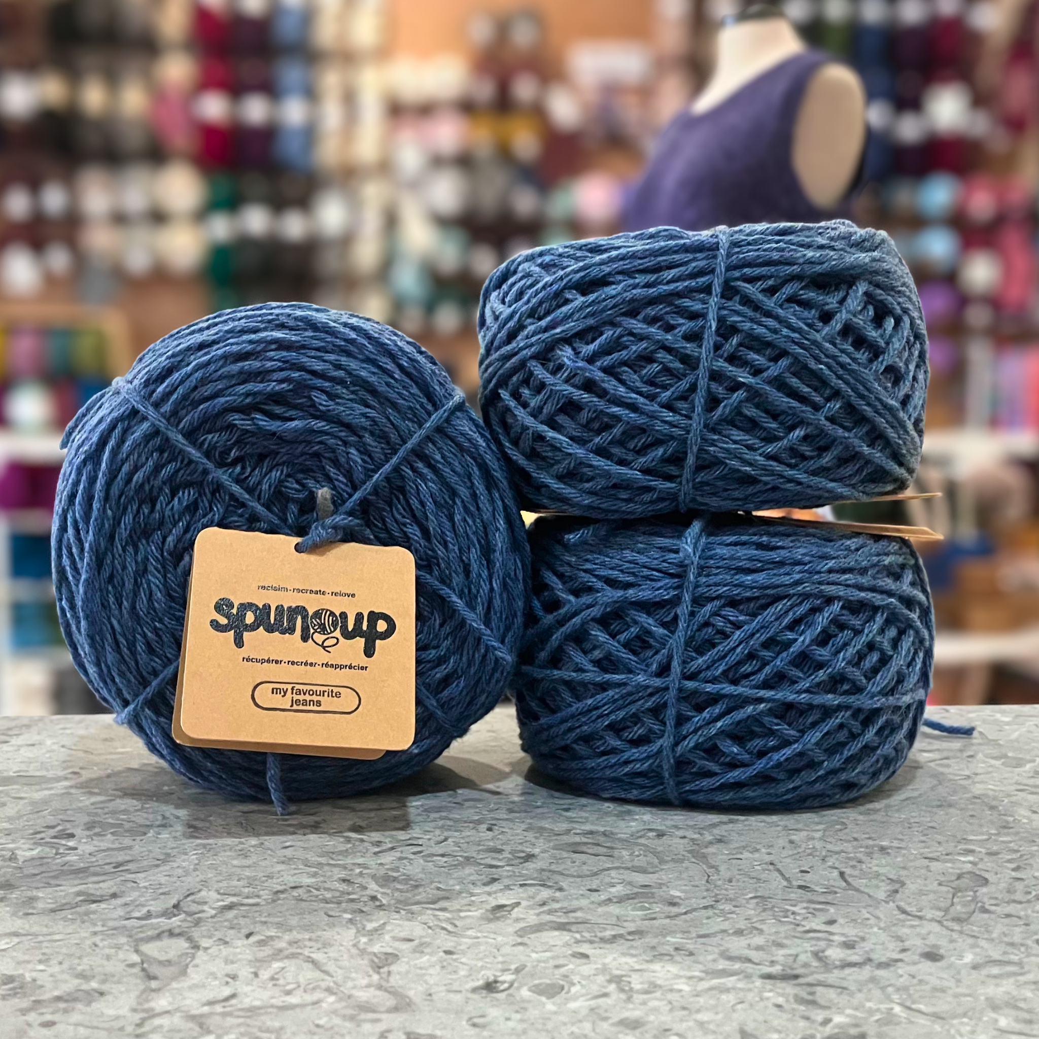 50g medium blue (my favourite jeans) GRS-certified recycled wool worsted yarn, 'Spun Up reclaim recreate relove' label visible, craft shop background (blurred)