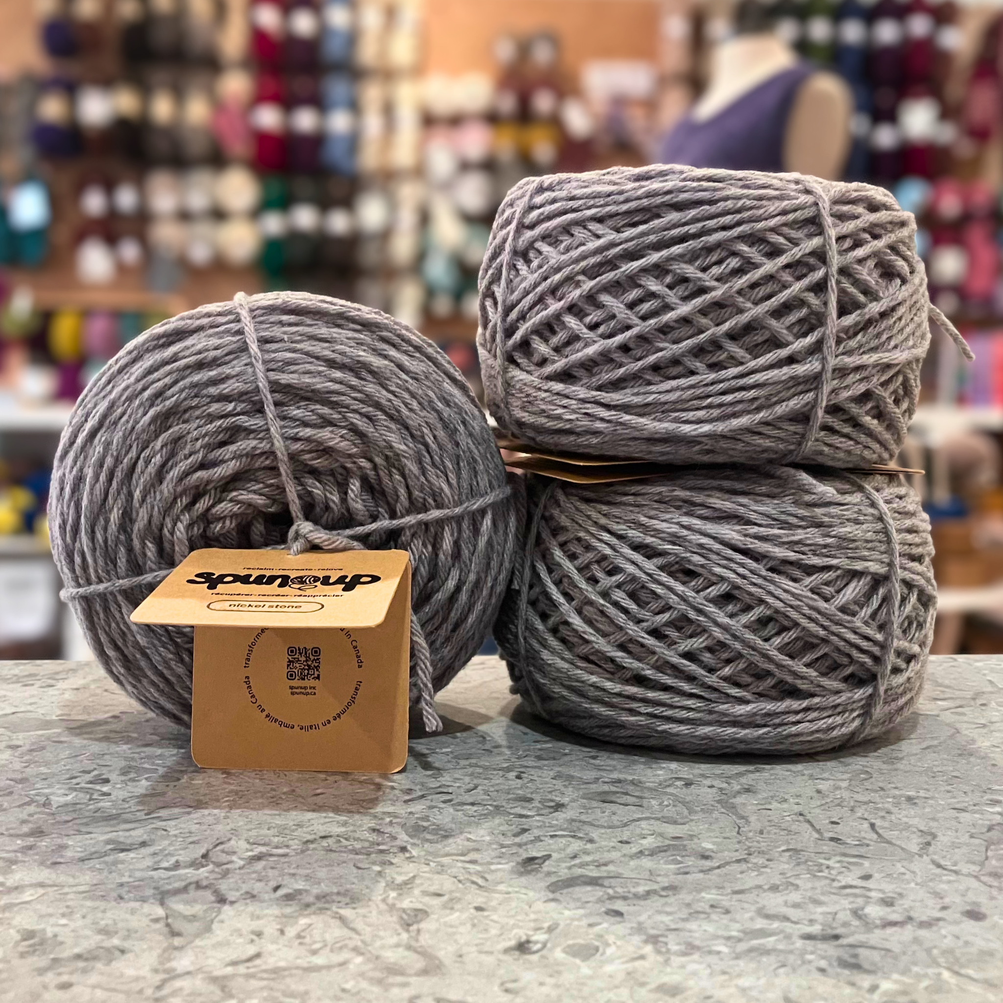 50g light grey (nickel stone) GRS-certified recycled wool worsted yarn, 'Spun Up reclaim recreate relove' label visible, craft shop background (blurred)