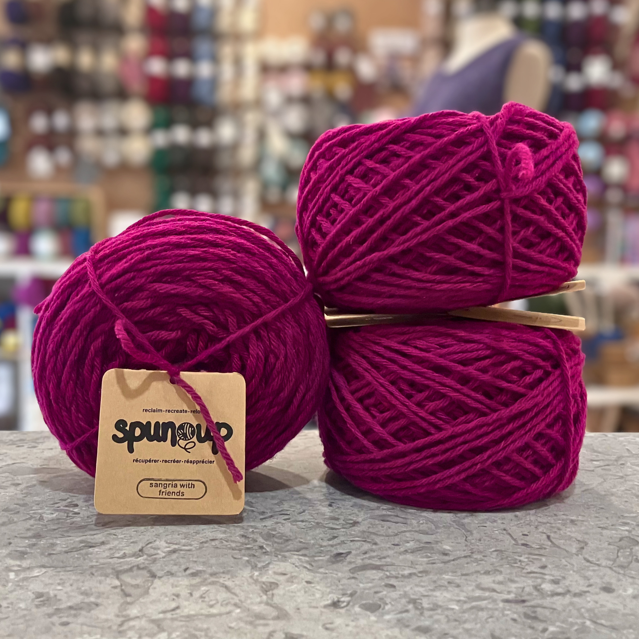 50g deep pink (sangria with friends) GRS-certified recycled wool worsted yarn, 'Spun Up reclaim recreate relove' label visible, craft shop background (blurred)