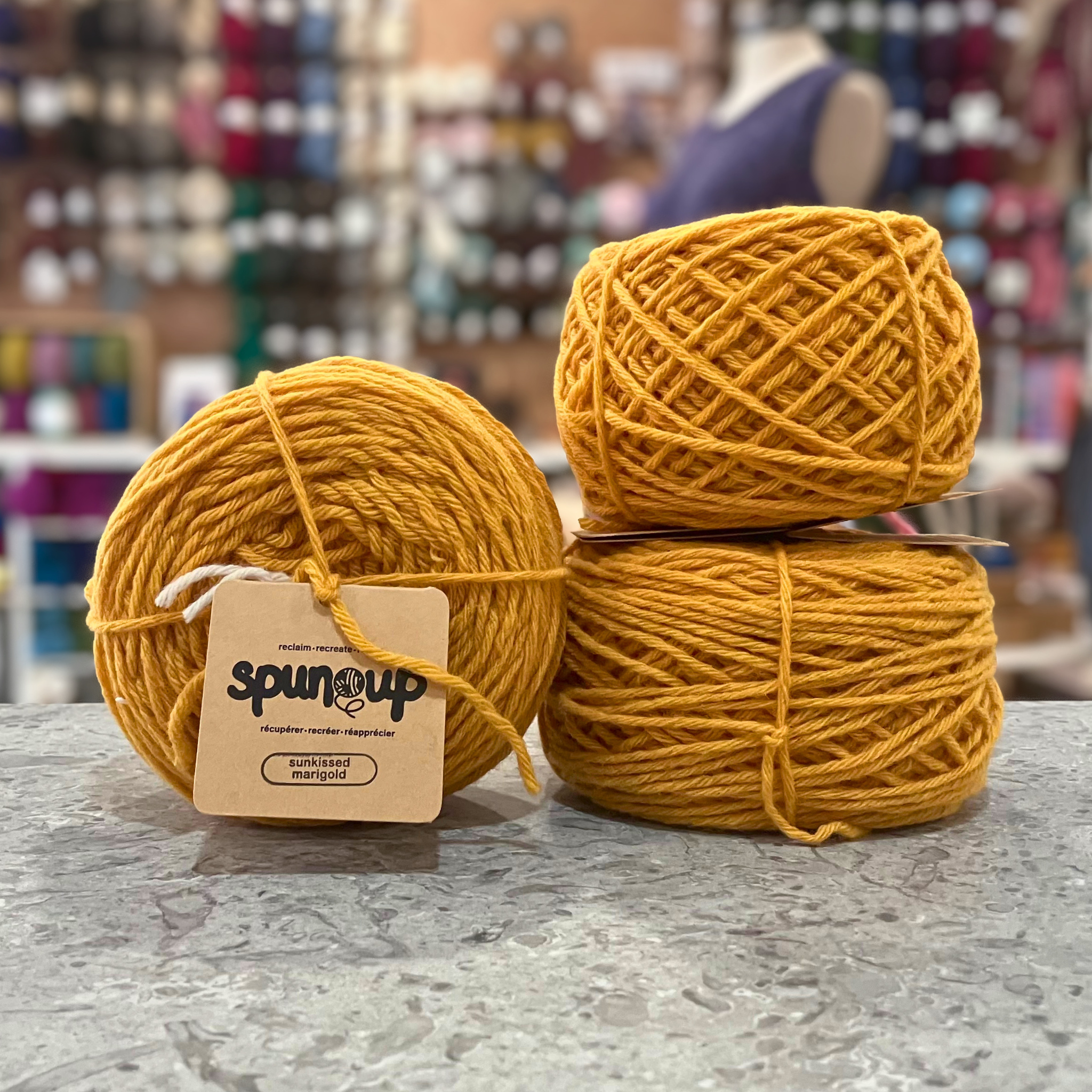50g bright yellow (sunkissed marigold) GRS-certified recycled wool worsted yarn, 'Spun Up reclaim recreate relove' label visible, craft shop background (blurred)