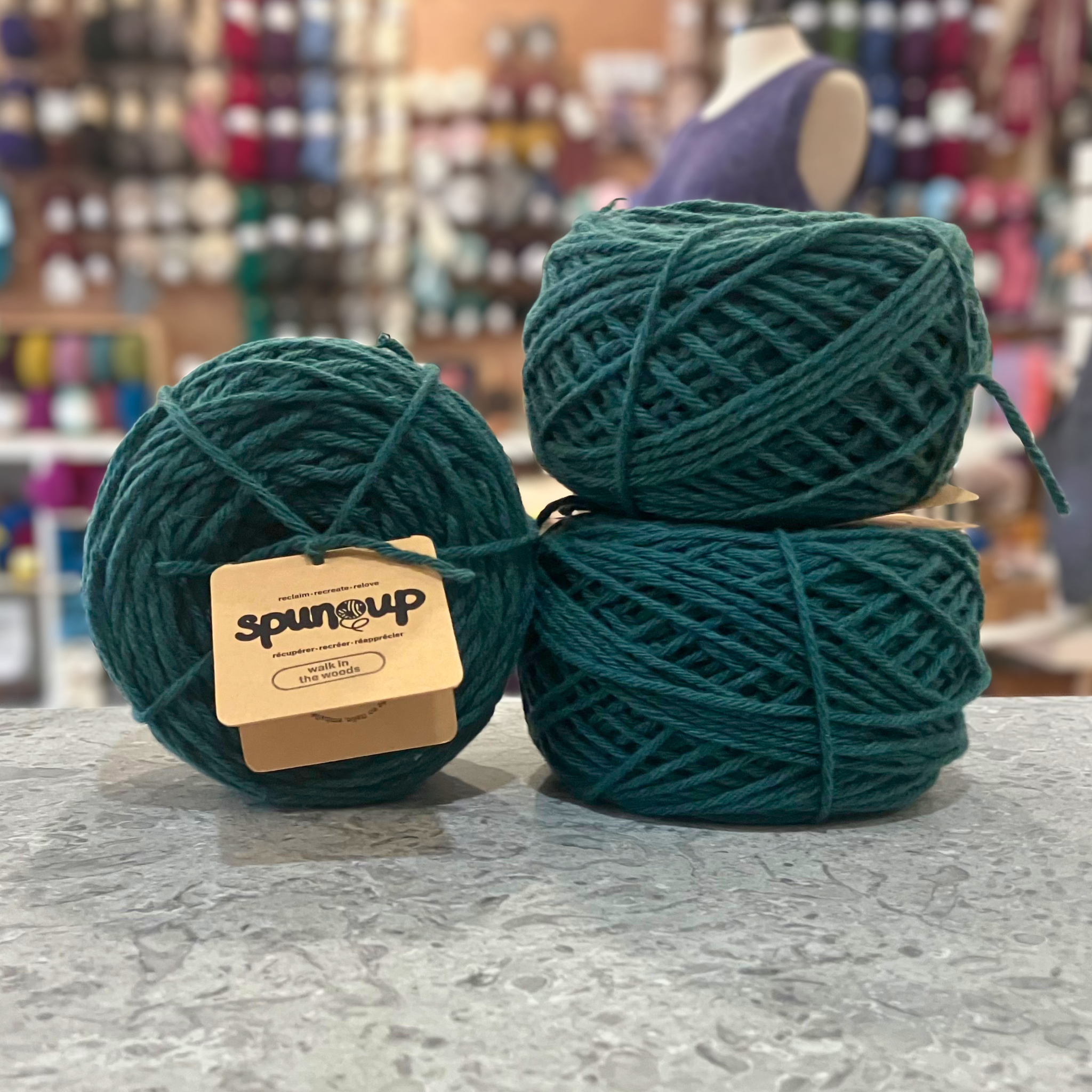 50g dark blue-green (walk in the woods) GRS-certified recycled wool worsted yarn, 'Spun Up reclaim recreate relove' label visible, craft shop background (blurred)