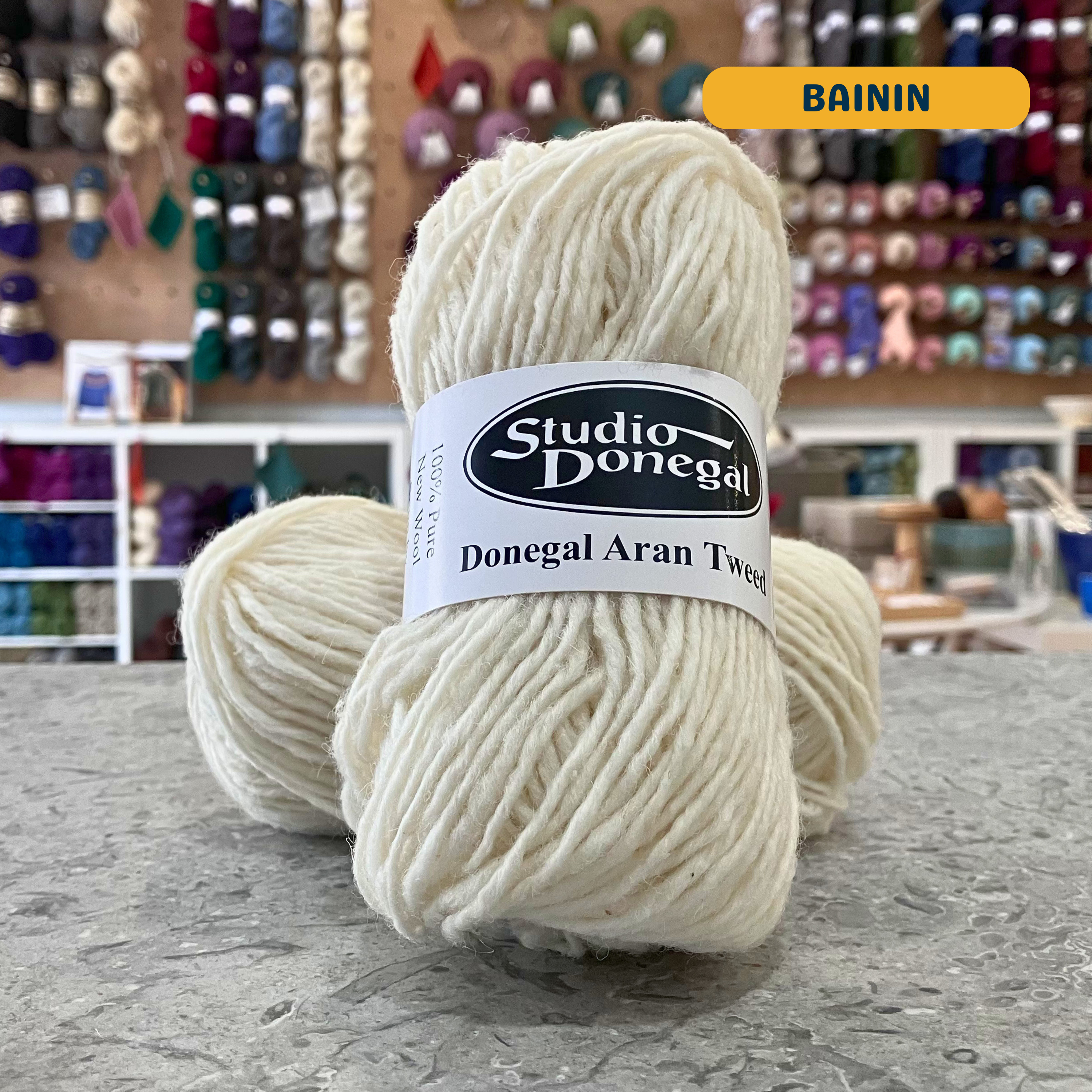50g natural white (bainin) Irish wool aran yarn, 'Studio Donegal Donegal Aran Tweed' label visible, craft shop background (blurred)
