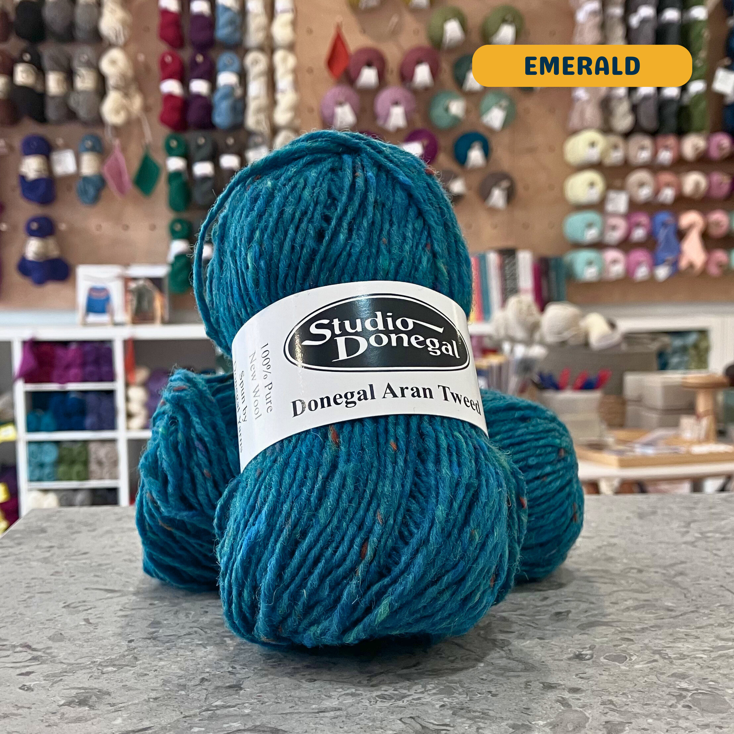 50g turquoise (emerald) with multi-coloured specks Irish wool aran yarn, 'Studio Donegal Donegal Aran Tweed' label visible, craft shop background (blurred)
