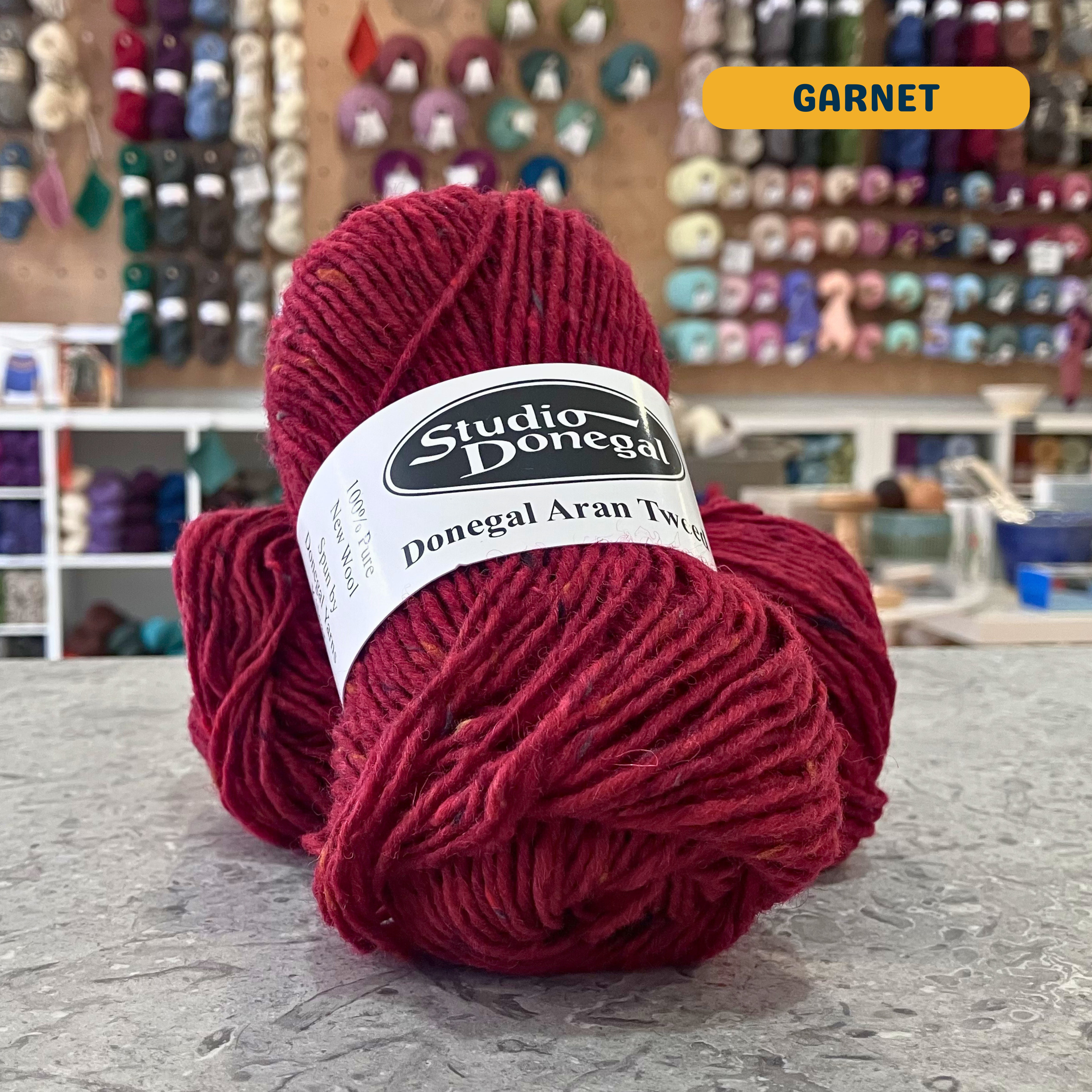 50g red (garnet) with multi-coloured specks Irish wool aran yarn, 'Studio Donegal Donegal Aran Tweed' label visible, craft shop background (blurred)