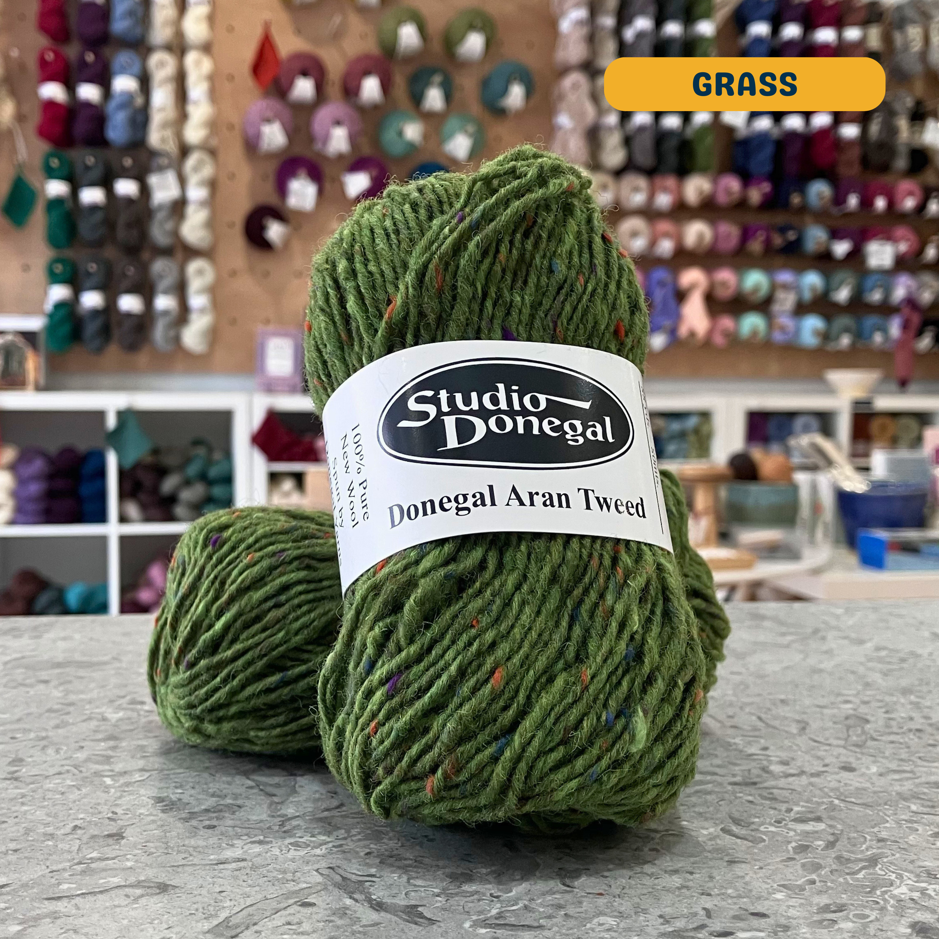 50g green (grass) with multi-coloured specks Irish wool aran yarn, 'Studio Donegal Donegal Aran Tweed' label visible, craft shop background (blurred)