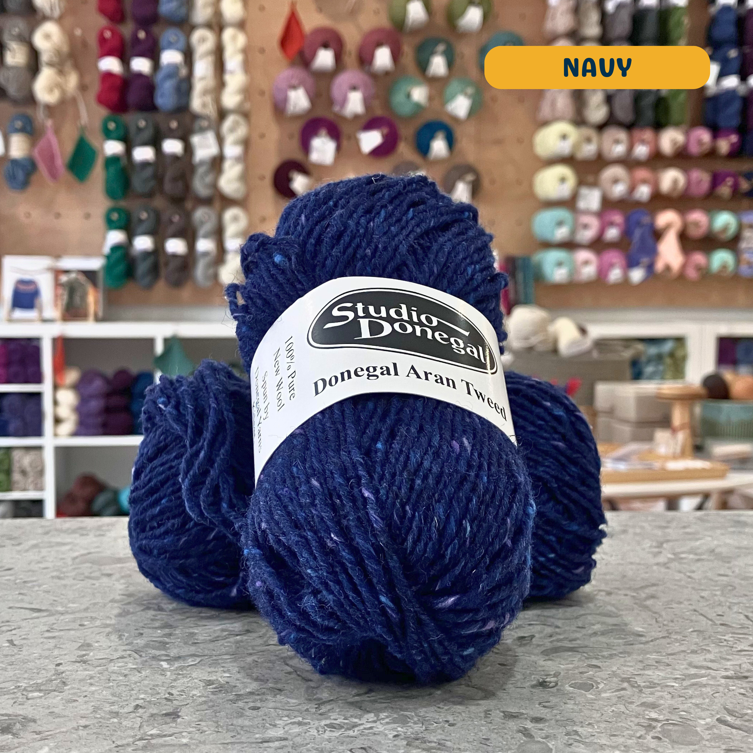 50g dark blue (navy) with multi-coloured specks Irish wool aran yarn, 'Studio Donegal Donegal Aran Tweed' label visible, craft shop background (blurred)