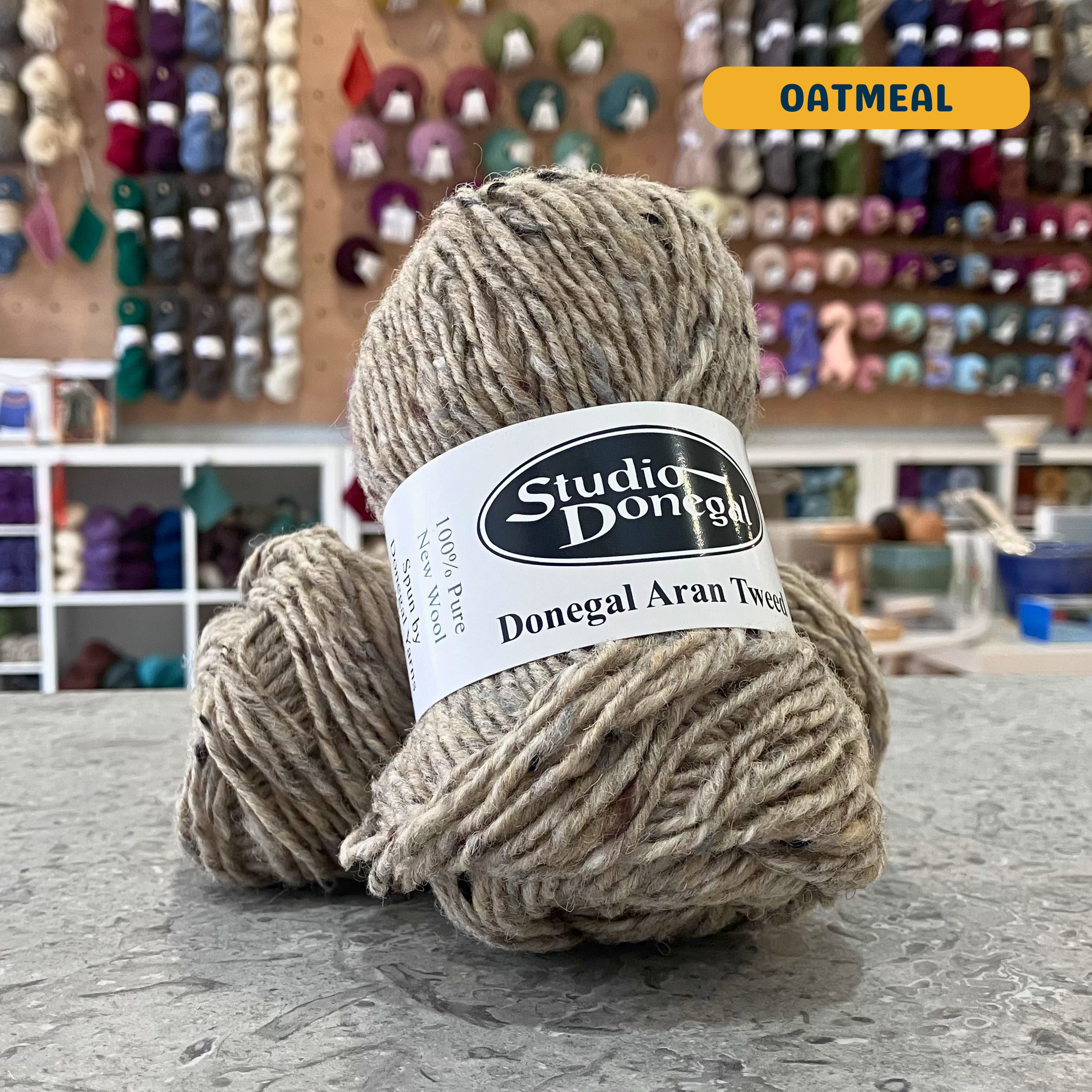 50g beige (oatmeal) with multi-coloured specks Irish wool aran yarn, 'Studio Donegal Donegal Aran Tweed' label visible, craft shop background (blurred)