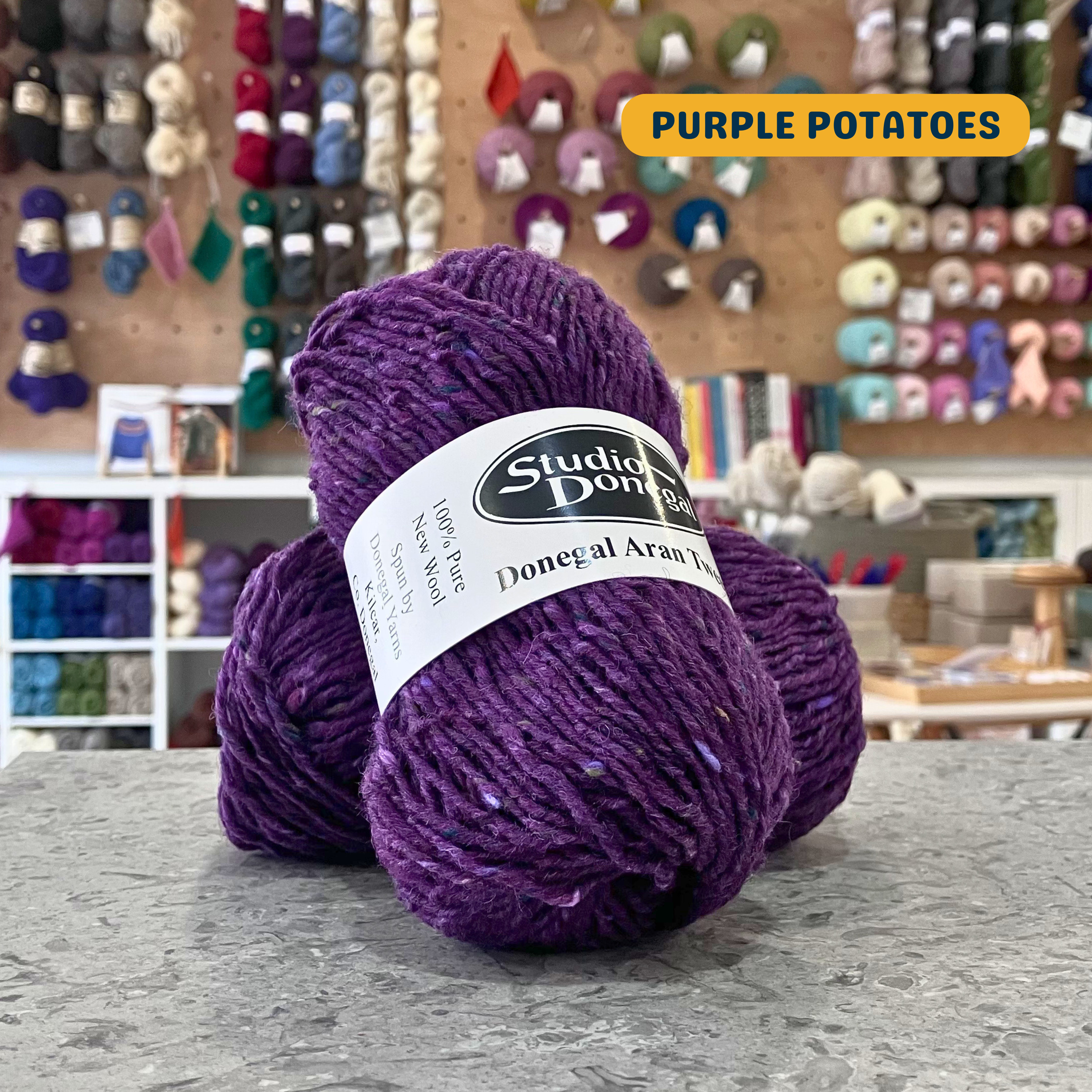 50g purple (purple potatoes) with multi-coloured specks Irish wool aran yarn, 'Studio Donegal Donegal Aran Tweed' label visible, craft shop background (blurred)