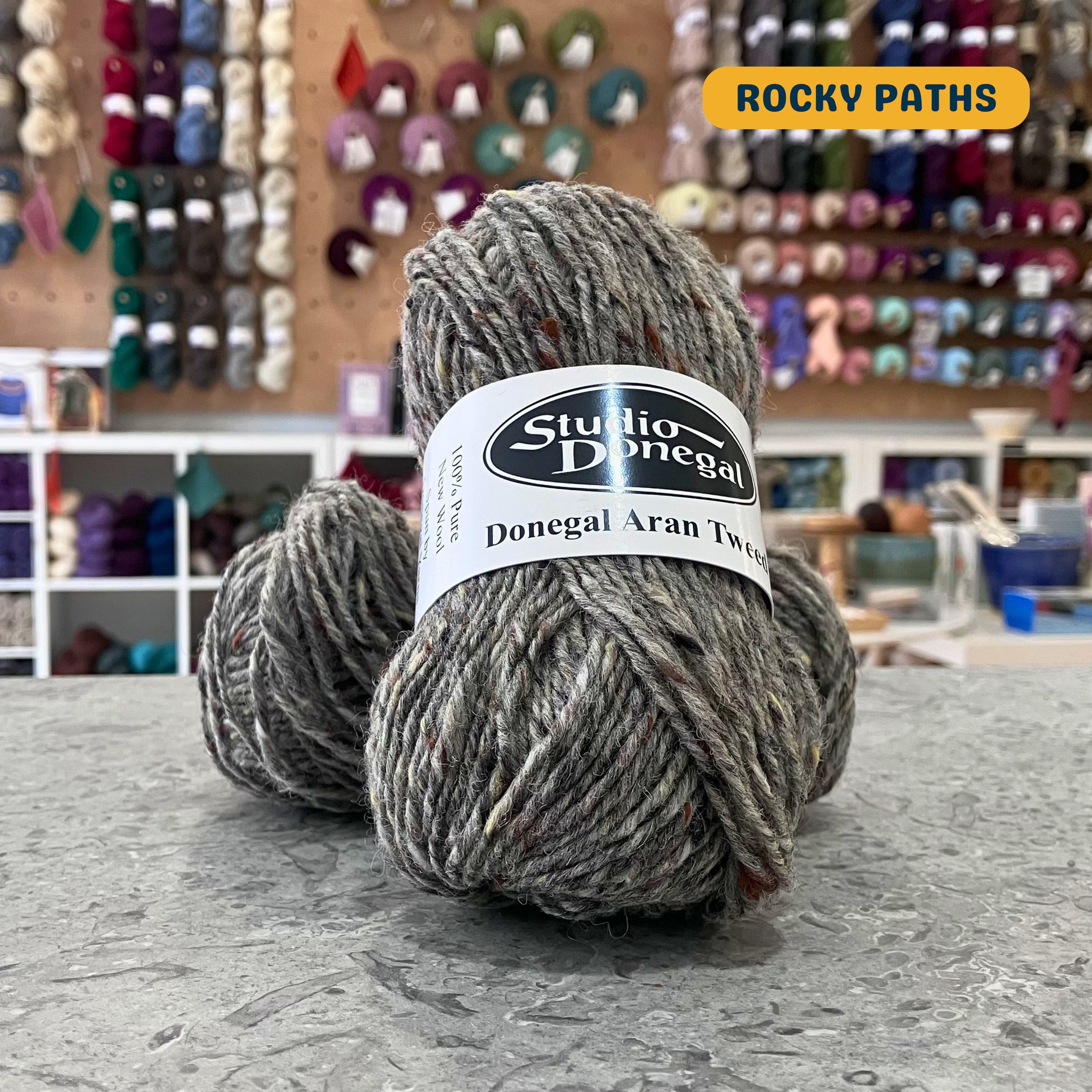 50g grey (rocky paths) with multi-coloured specks Irish wool aran yarn, 'Studio Donegal Donegal Aran Tweed' label visible, craft shop background (blurred)
