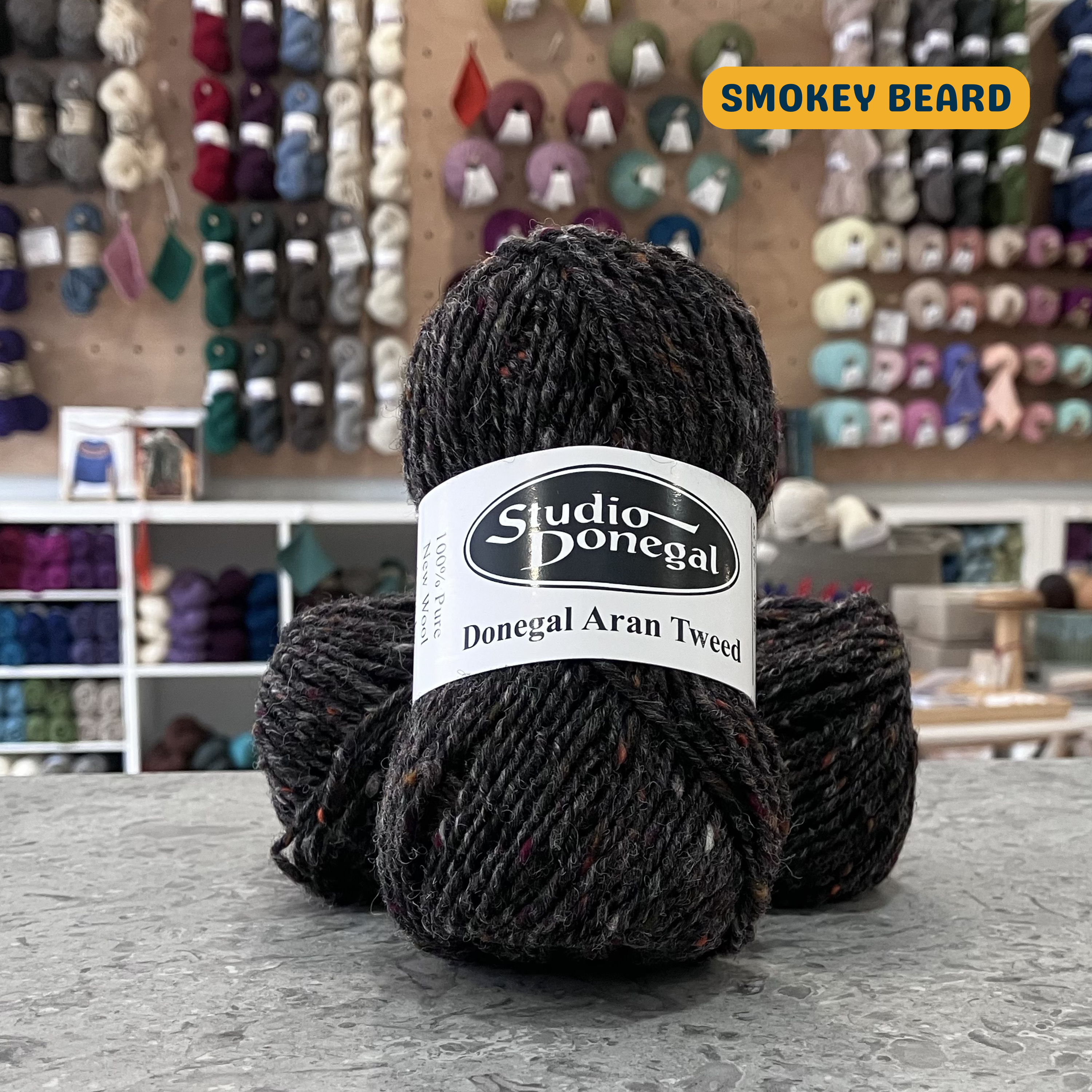 50g dark grey (smokey beard) with multi-coloured specks Irish wool aran yarn, 'Studio Donegal Donegal Aran Tweed' label visible, craft shop background (blurred)