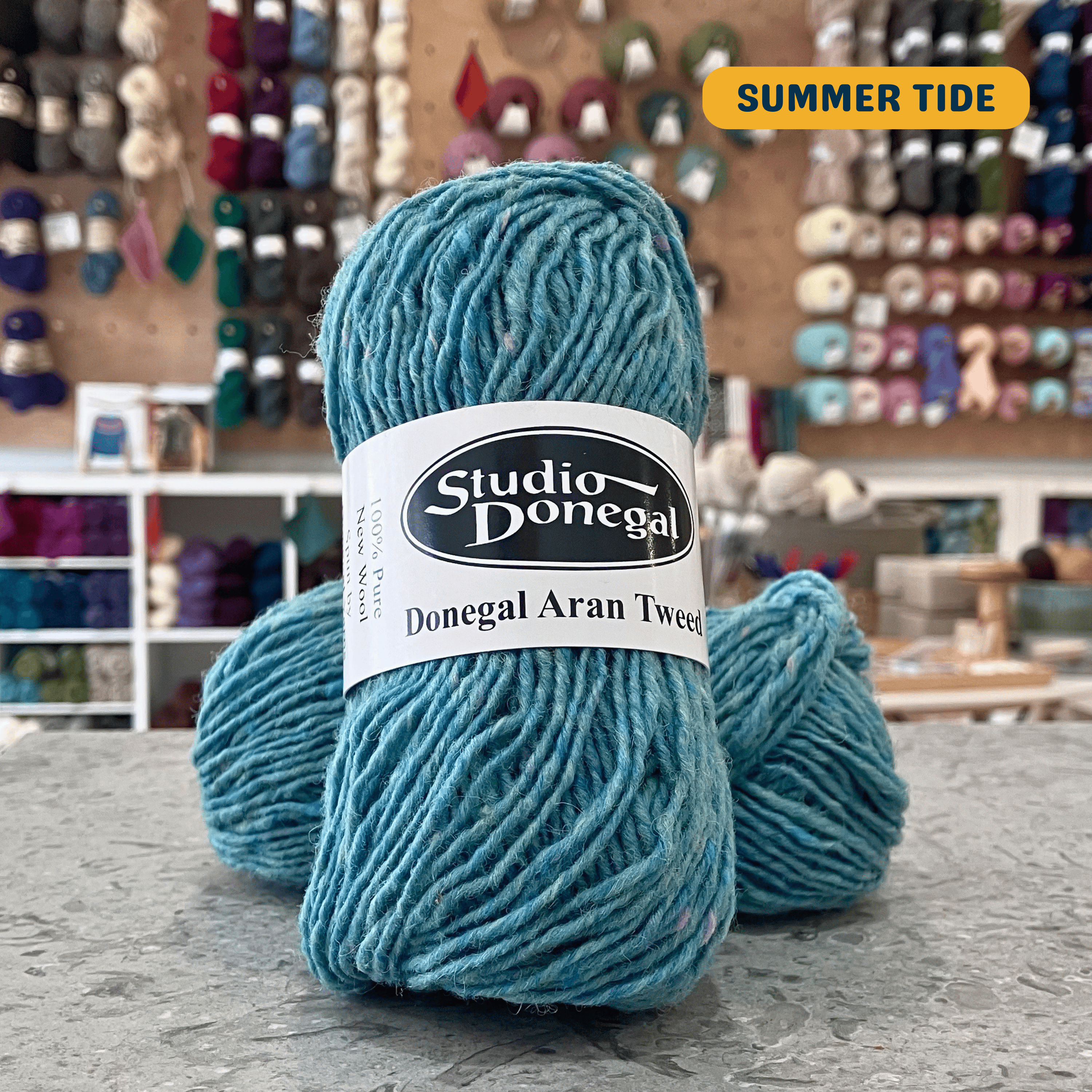 50g light turquoise (summer tide) with multi-coloured specks Irish wool aran yarn, 'Studio Donegal Donegal Aran Tweed' label visible, craft shop background (blurred)